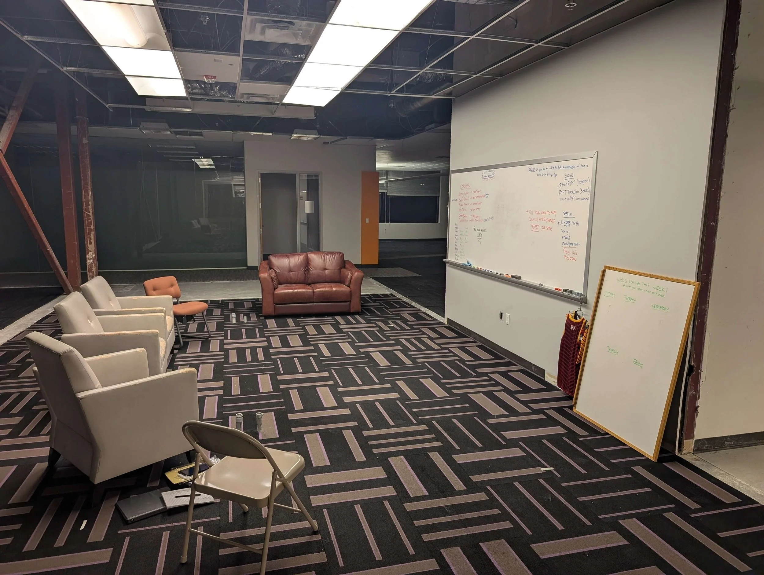 An office or conference room with chairs and a whiteboard. There are five chairs, a leather couch, and a small table with a notebook on it. The room has modern lighting fixtures and a patterned carpet. A whiteboard leans against the wall near a parti