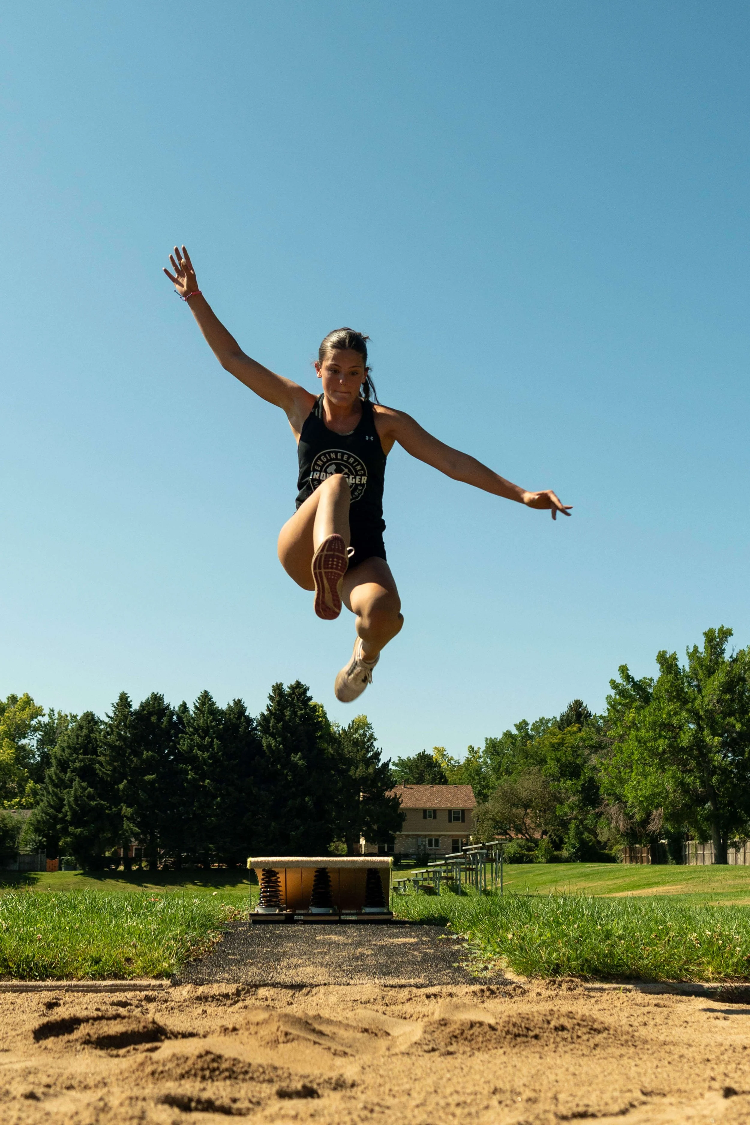 A female long jumper mid-air during a jump at an outdoor track and field event, with a clear blue sky and trees in the background.