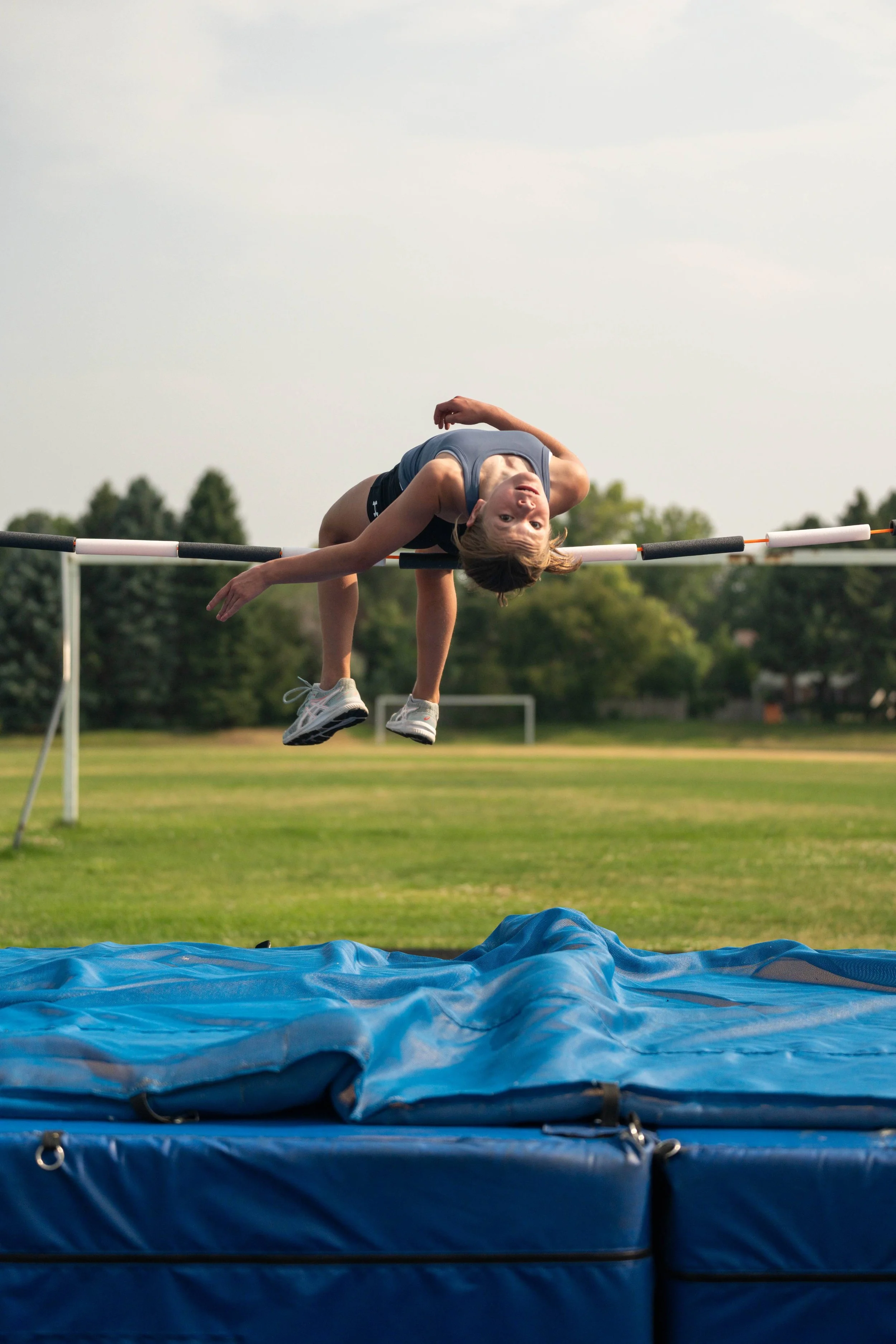A young girl in athletic clothing attempting to clear a high jump bar during a track and field practice or competition on an outdoor field.
