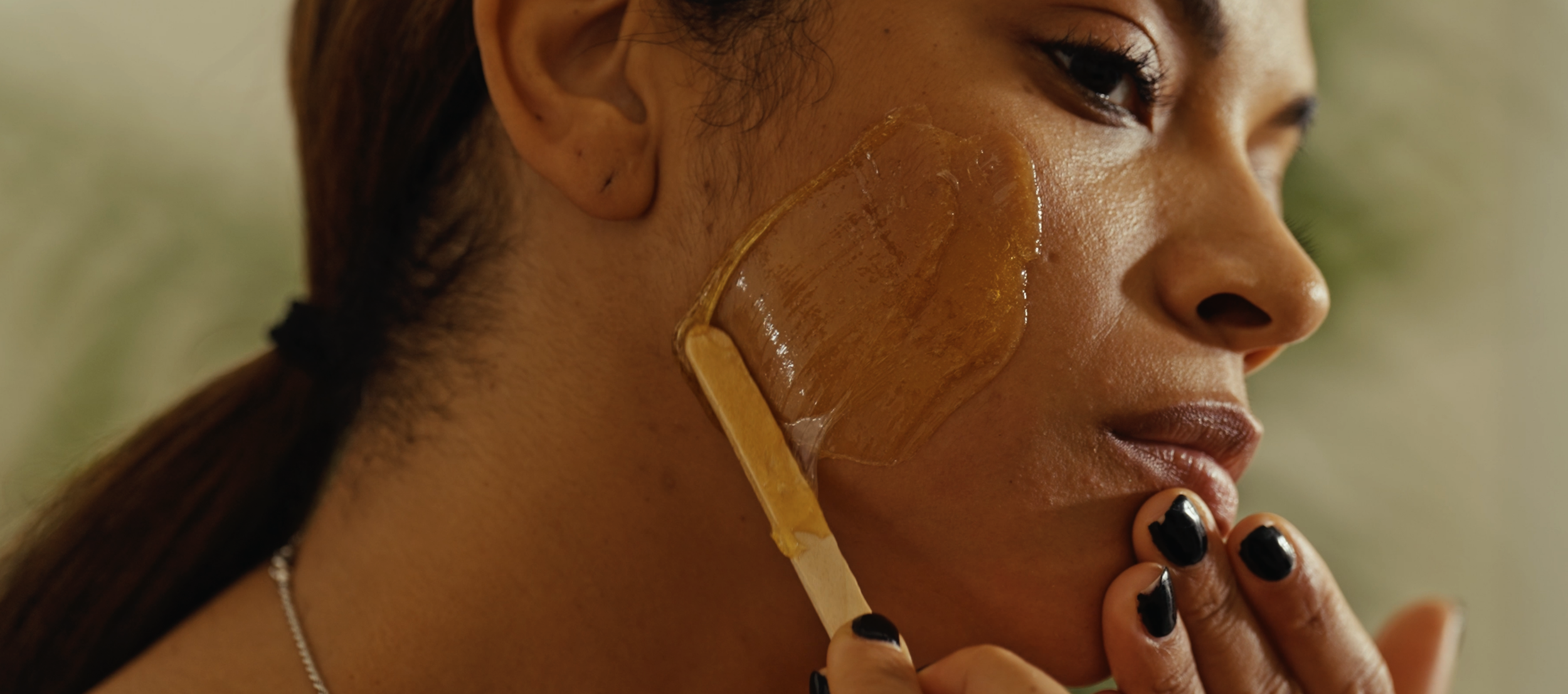 A woman with dark hair tied in a ponytail, applying a transparent, brownish skincare mask to her cheek with a spatula, with her fingers near her chin. She is outdoors with a blurred background.