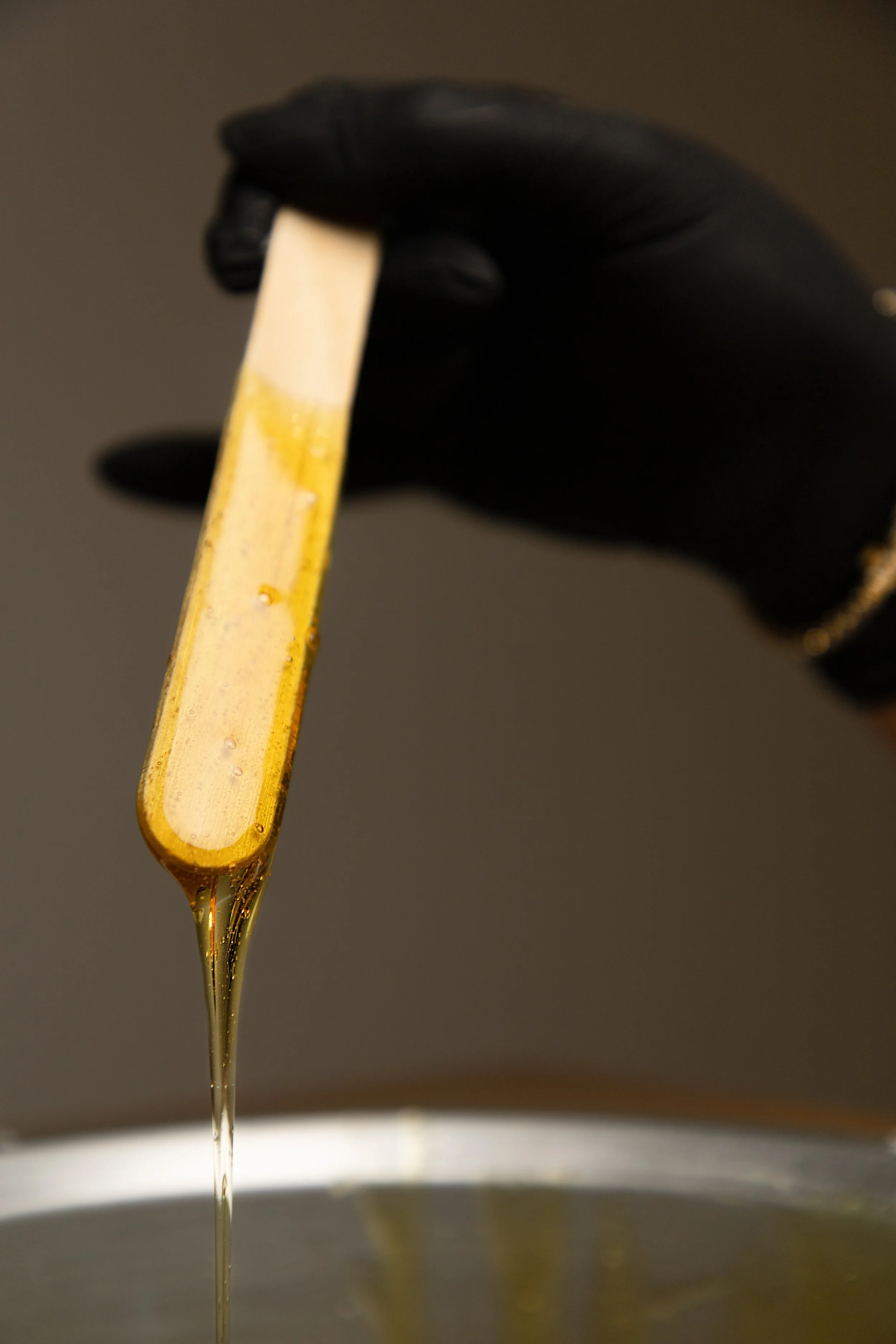 Close-up of a black-gloved hand holding a wooden stick coated with honey, with honey dripping from the stick into a container below.