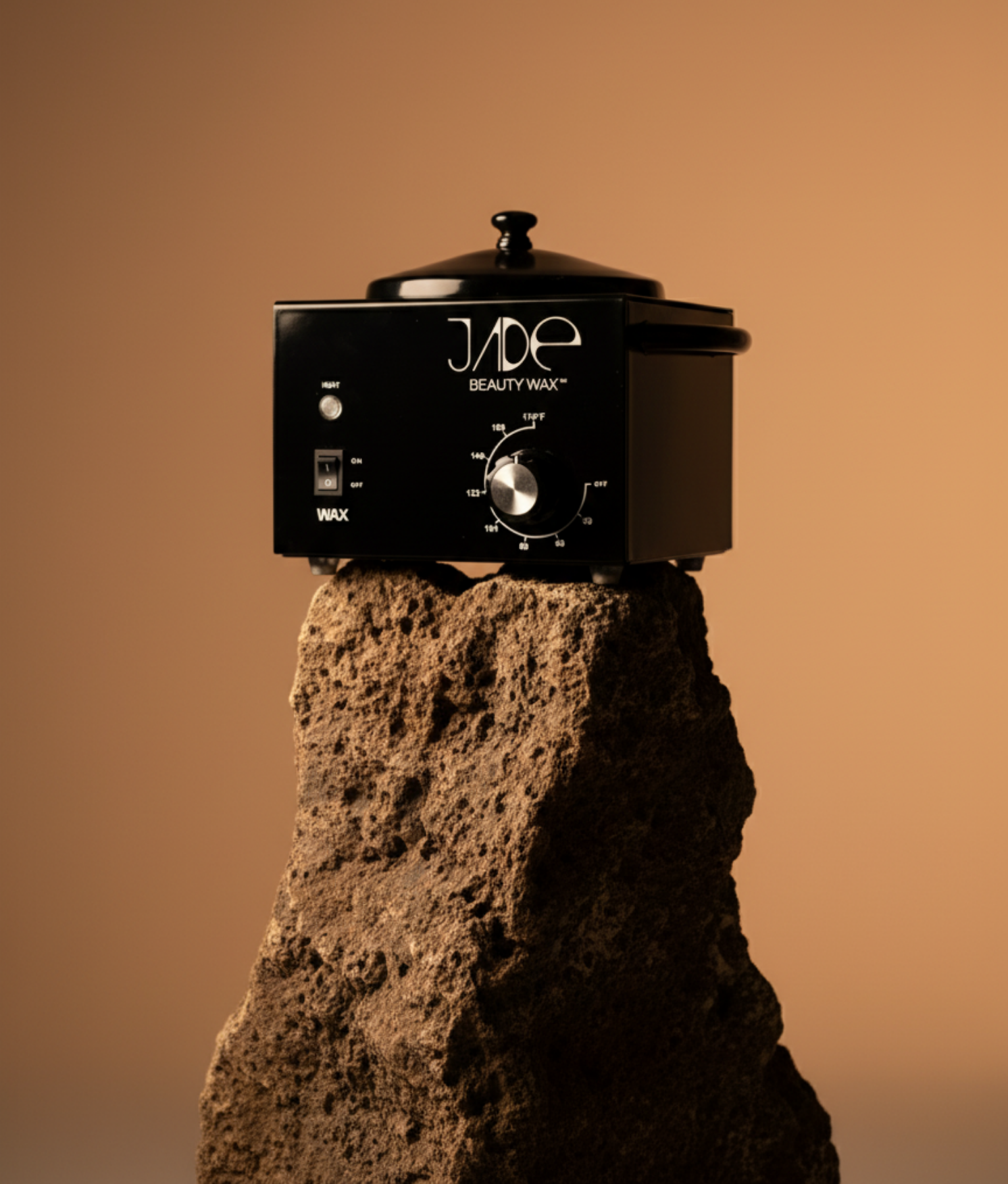 A black wax warmer labeled 'Jade Beauty Wax' is placed on top of a large, textured rock with a neutral background.