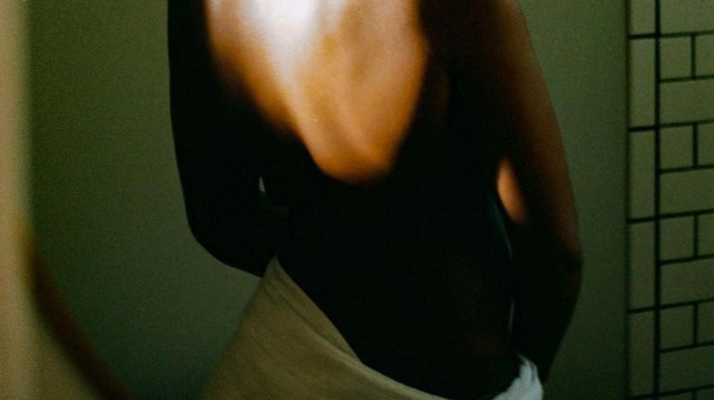 Close-up of a person bending forward, wearing a black top with a deep V neckline and a tan skirt, standing in front of a mirror reflecting part of a room with a brick wall.