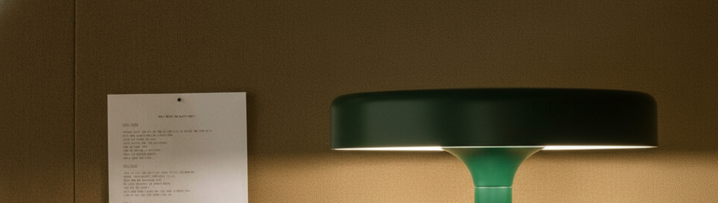 A green desk lamp on a wooden surface with a wall and a printed sheet of paper in the background.