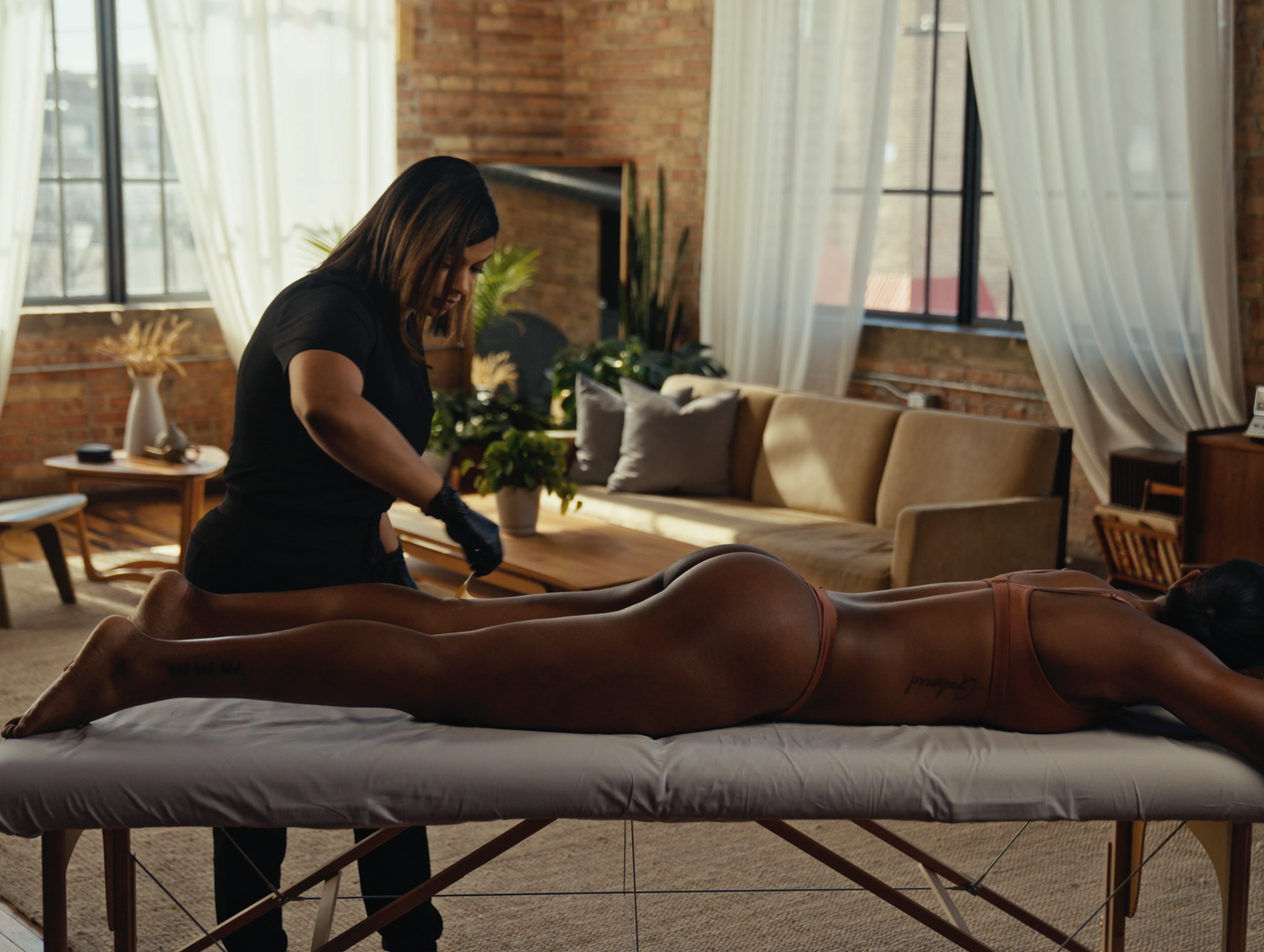 A woman receiving a massage on a massage table in a cozy living room with brick walls, large windows, and white curtains.