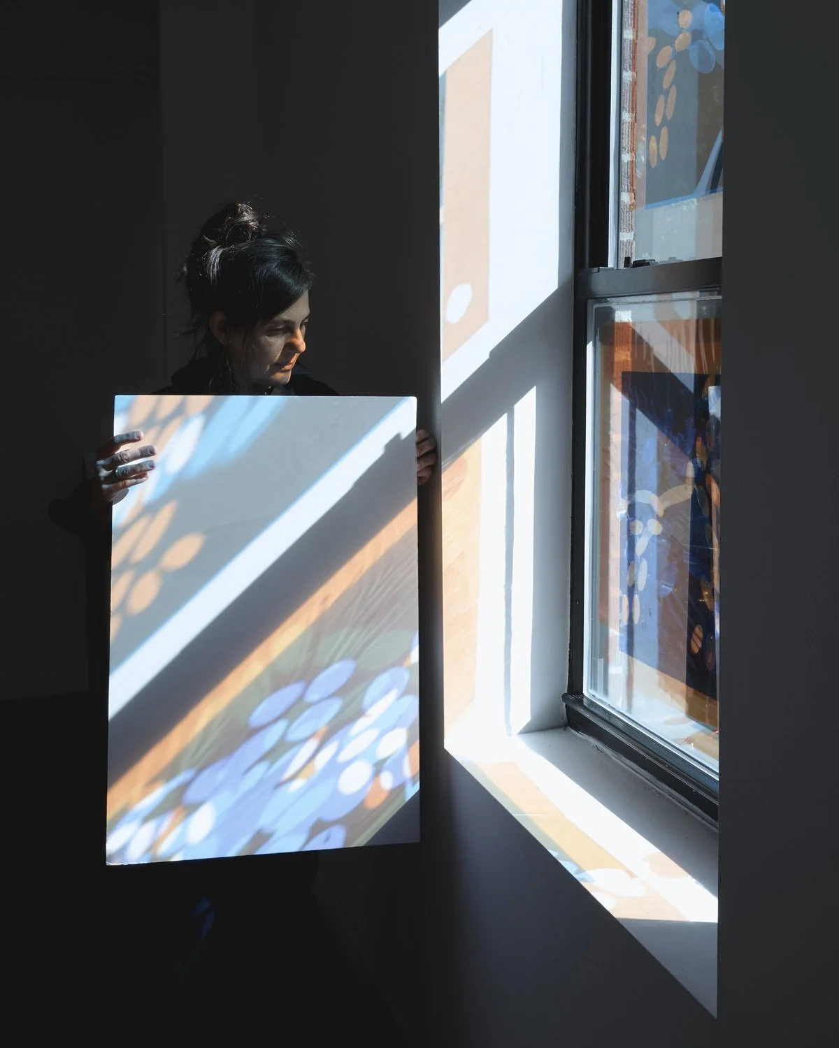 Photo of artist Lia Zuvilivia holding a white board that catches a colorful projection of light from a nearby window.