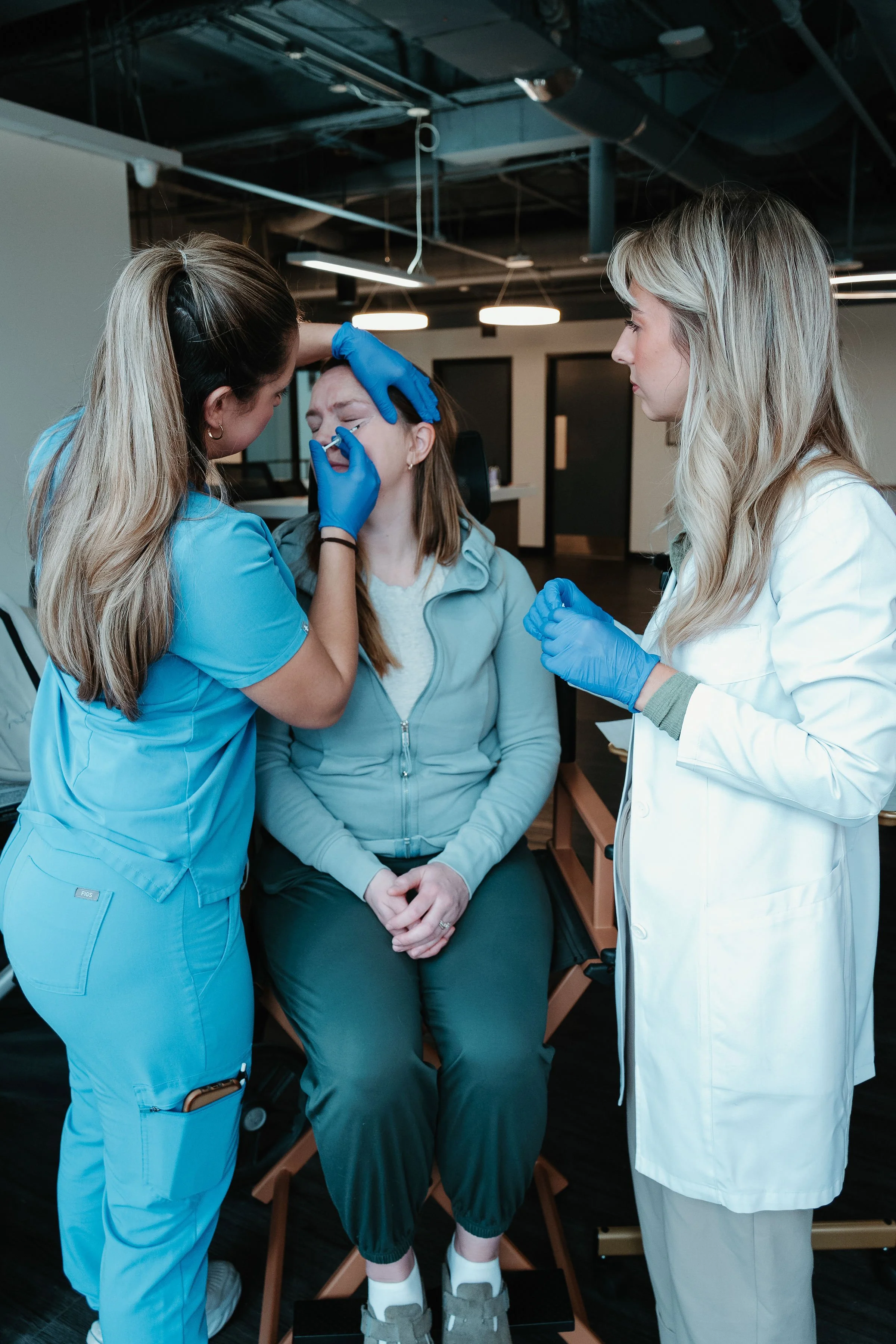 Two healthcare professionals perform a medical procedure on a seated woman in a clinical setting.