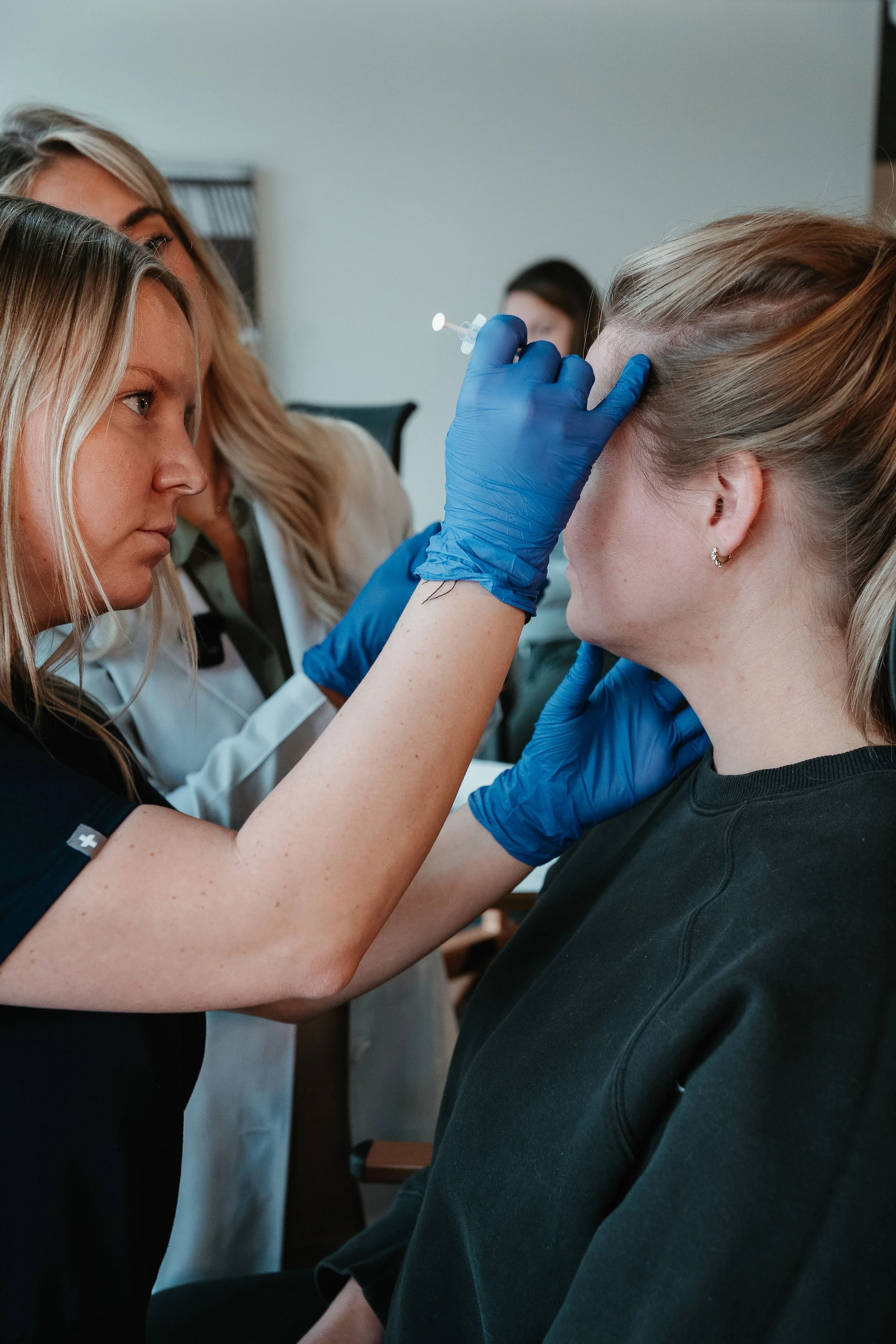 A healthcare professional administering a vaccine to a woman in a black shirt, with another healthcare worker and a person in the background.