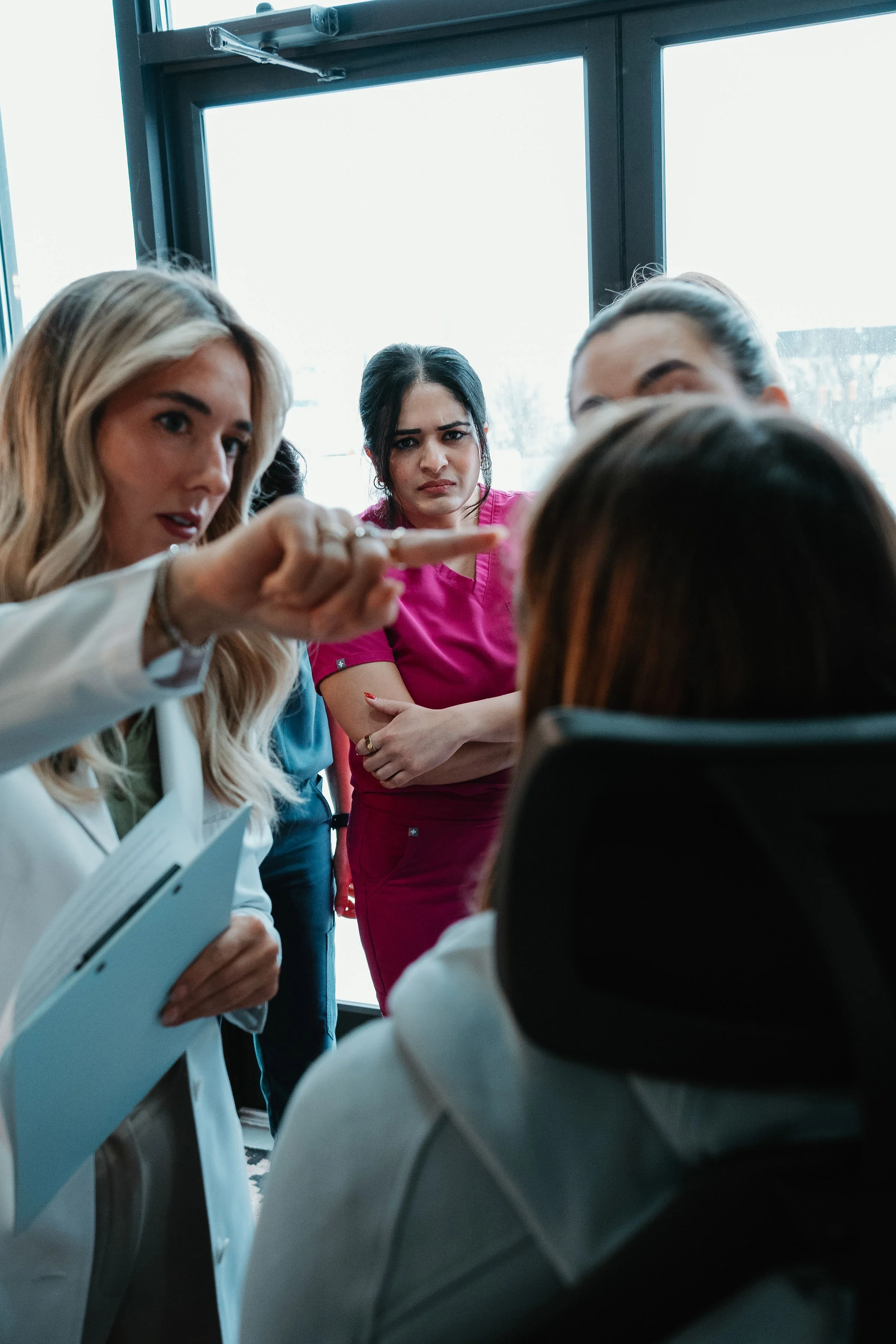 A woman in a white coat is pointing and speaking to a group of people, including a woman in a pink shirt with crossed arms and a woman with brown hair sitting, during a discussion in a room with large windows.