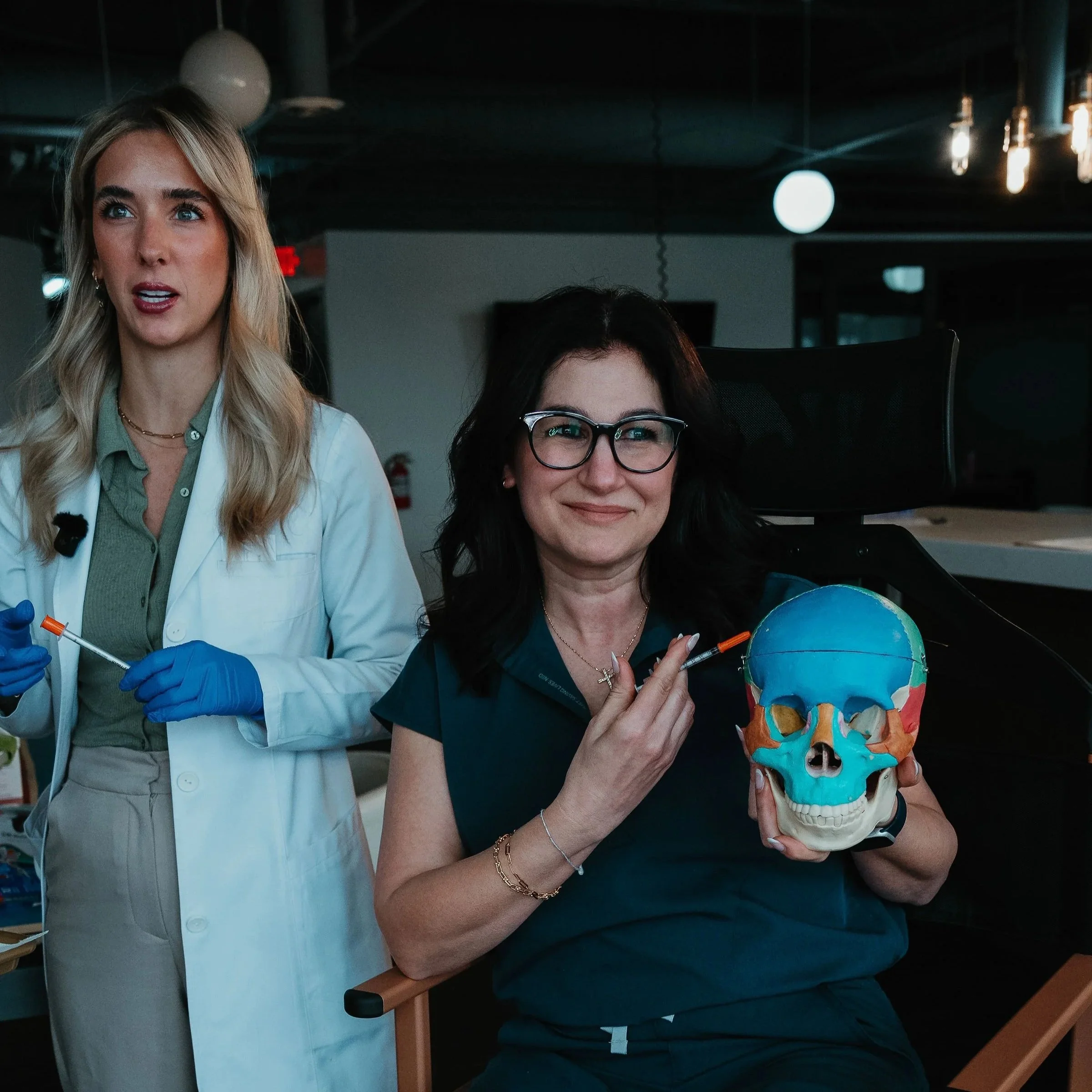 A woman with dark hair and glasses holding a painted skull with bright colors in an indoor setting, with another woman in a white coat and blue gloves standing beside her.