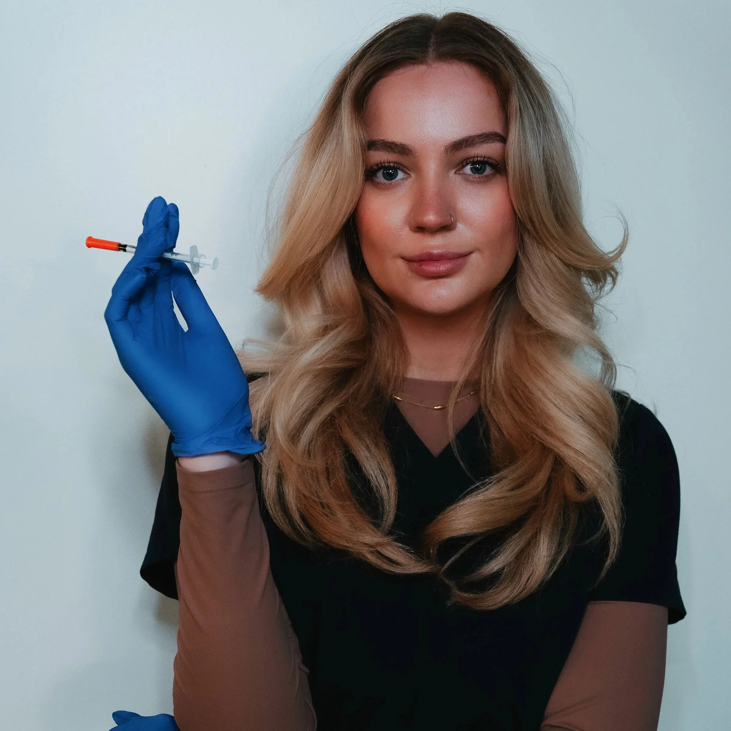 A woman with long blonde hair wearing a black shirt, holding a syringe with a blue glove, ready for vaccination.