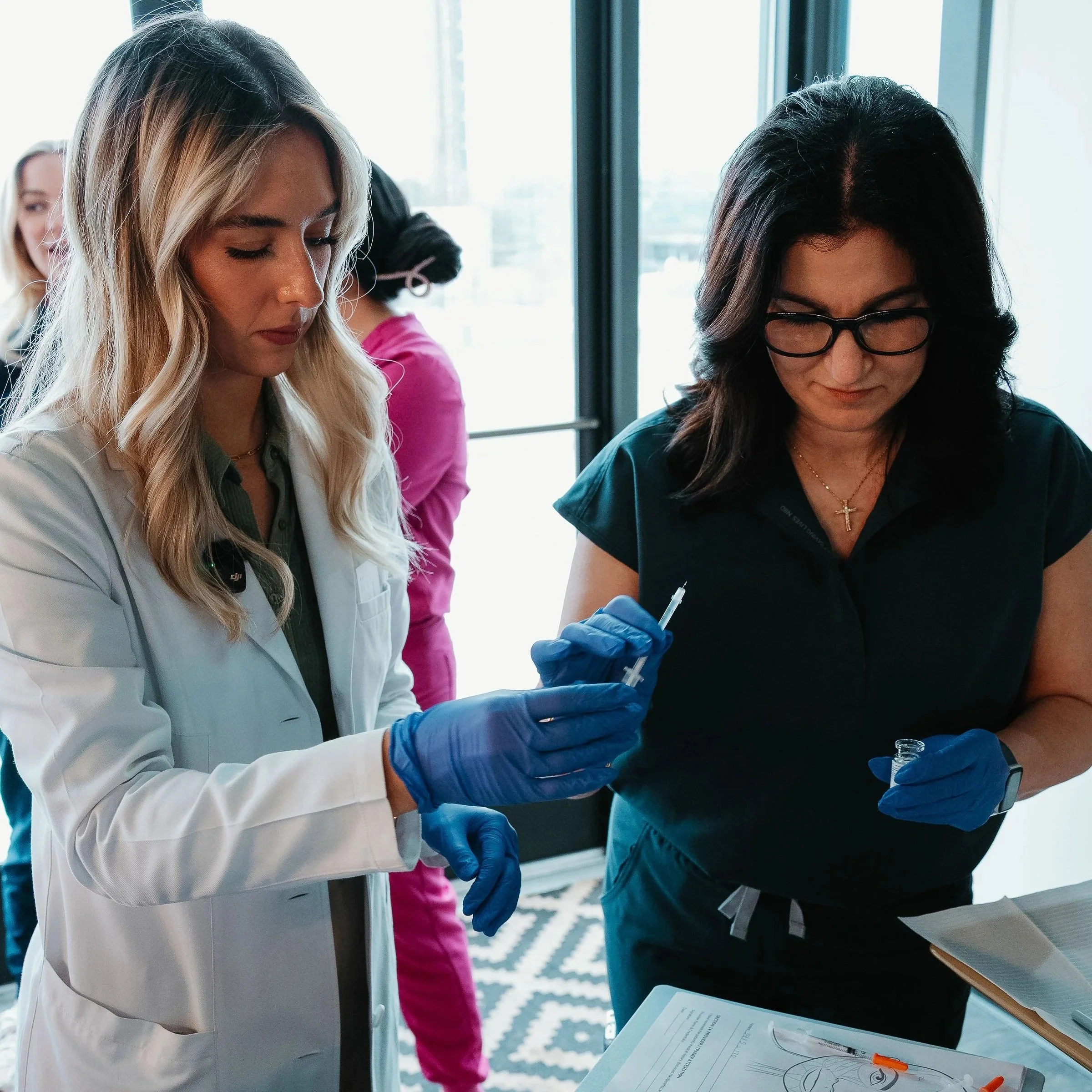 Two women wearing gloves and holding a syringe in a room with large windows.