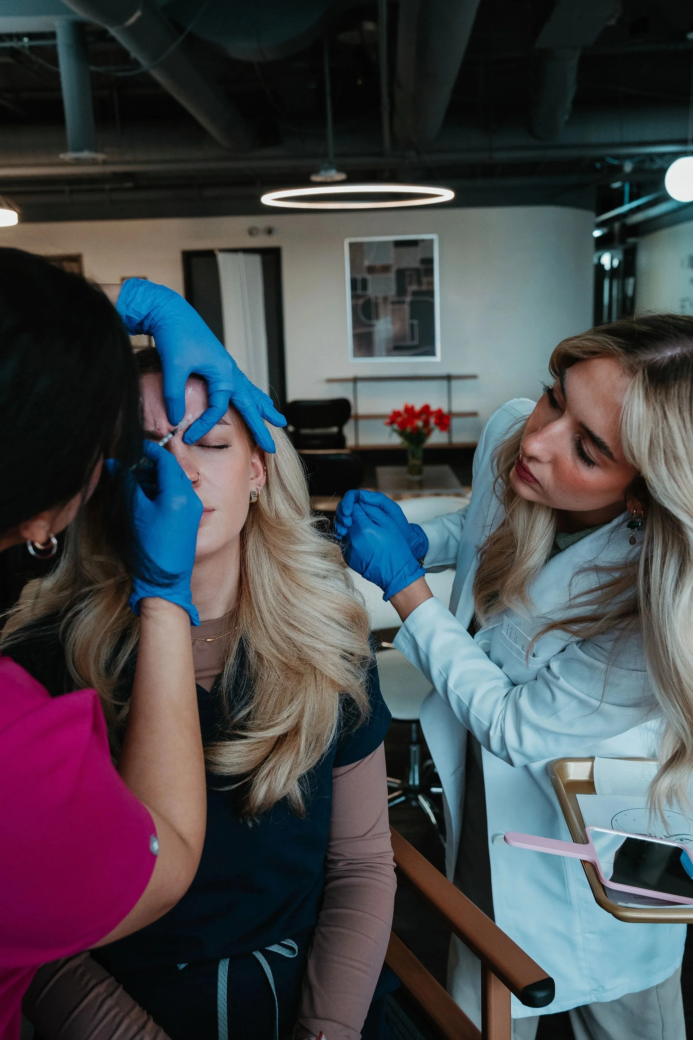Two women, one in a white coat, provide cosmetic treatment to a woman with blonde hair seated in a chair inside a modern clinic.