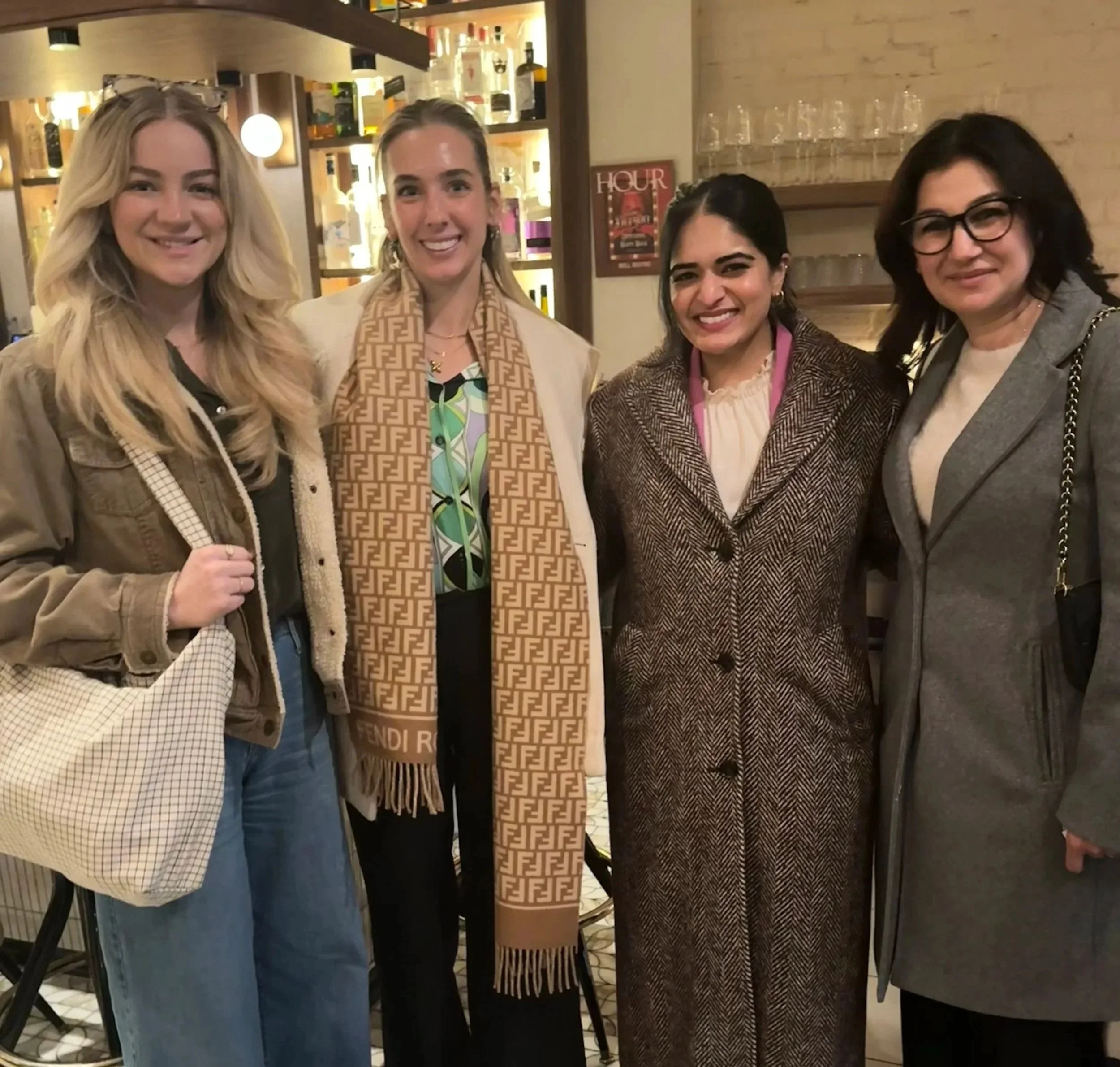 Four women standing together inside a restaurant or bar, smiling at the camera, with a decorated shelf and glasses in the background.
