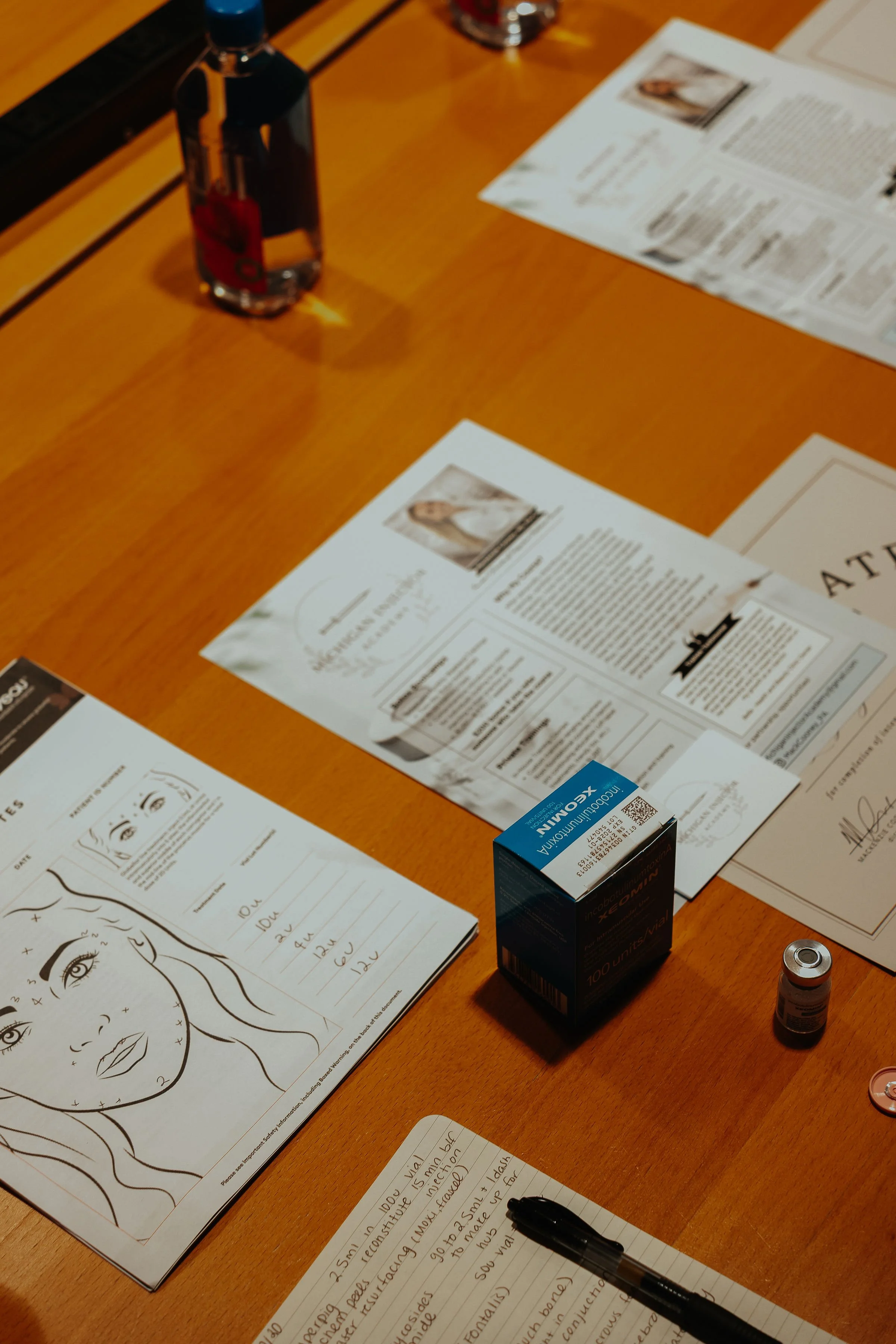 A wooden table with brochures, a notebook with handwritten notes, a pen, a bottle of eye drops, and a small medication container.