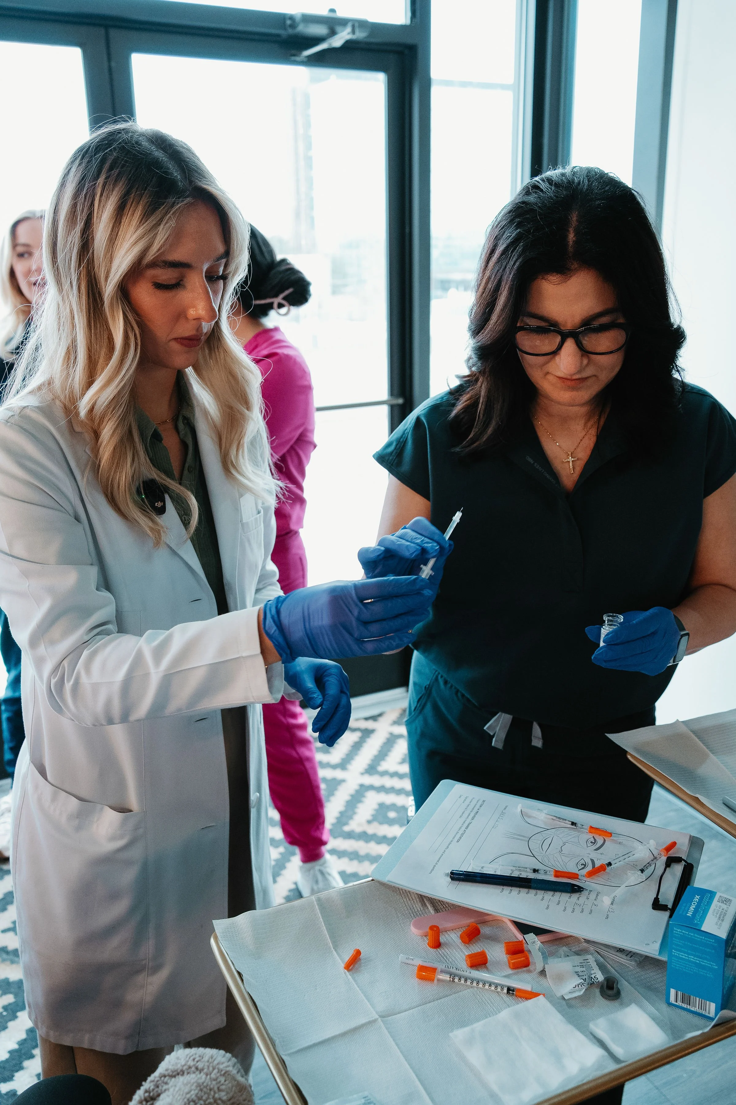 A healthcare professional, wearing a white coat and blue gloves, prepares a syringe, while another woman with glasses, also wearing blue gloves, watches. There are medical supplies and papers on a table in front of them.