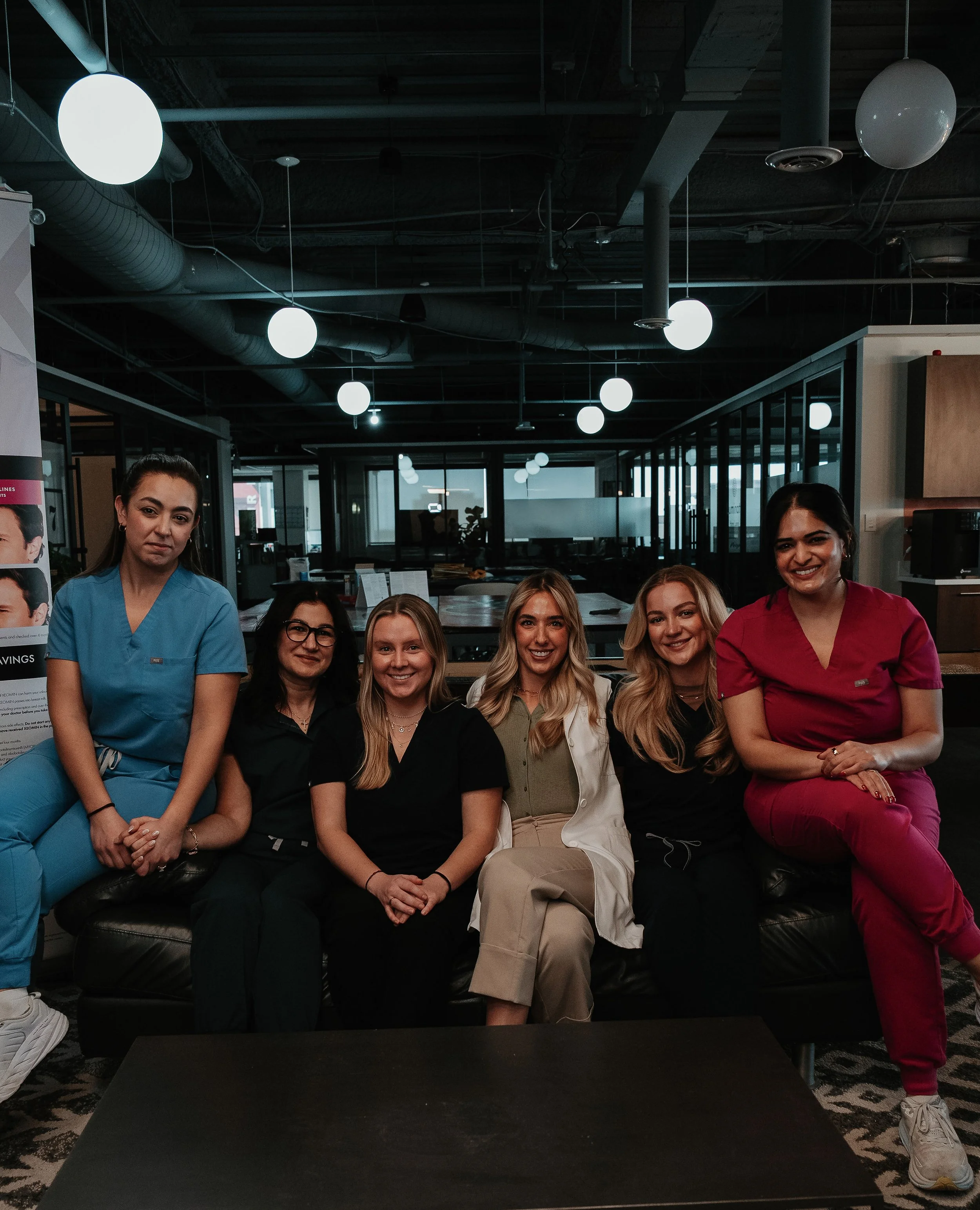 Six women sitting and standing on a black sofa in an indoor setting, some wearing scrubs and others in casual clothing, with a modern office or lounge background featuring glass partitions and round ceiling lights.