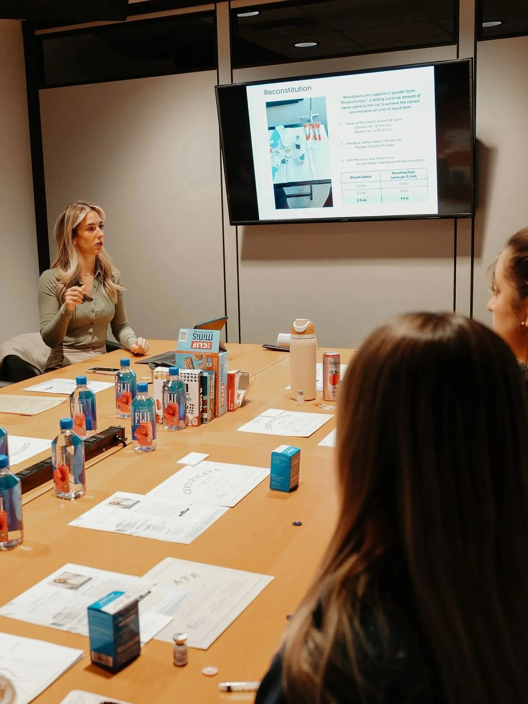 A woman is giving a presentation in a conference room, with a slide titled 'Reconstitution' displayed on a large screen. The table in front of her has water bottles, papers, and medical supplies.