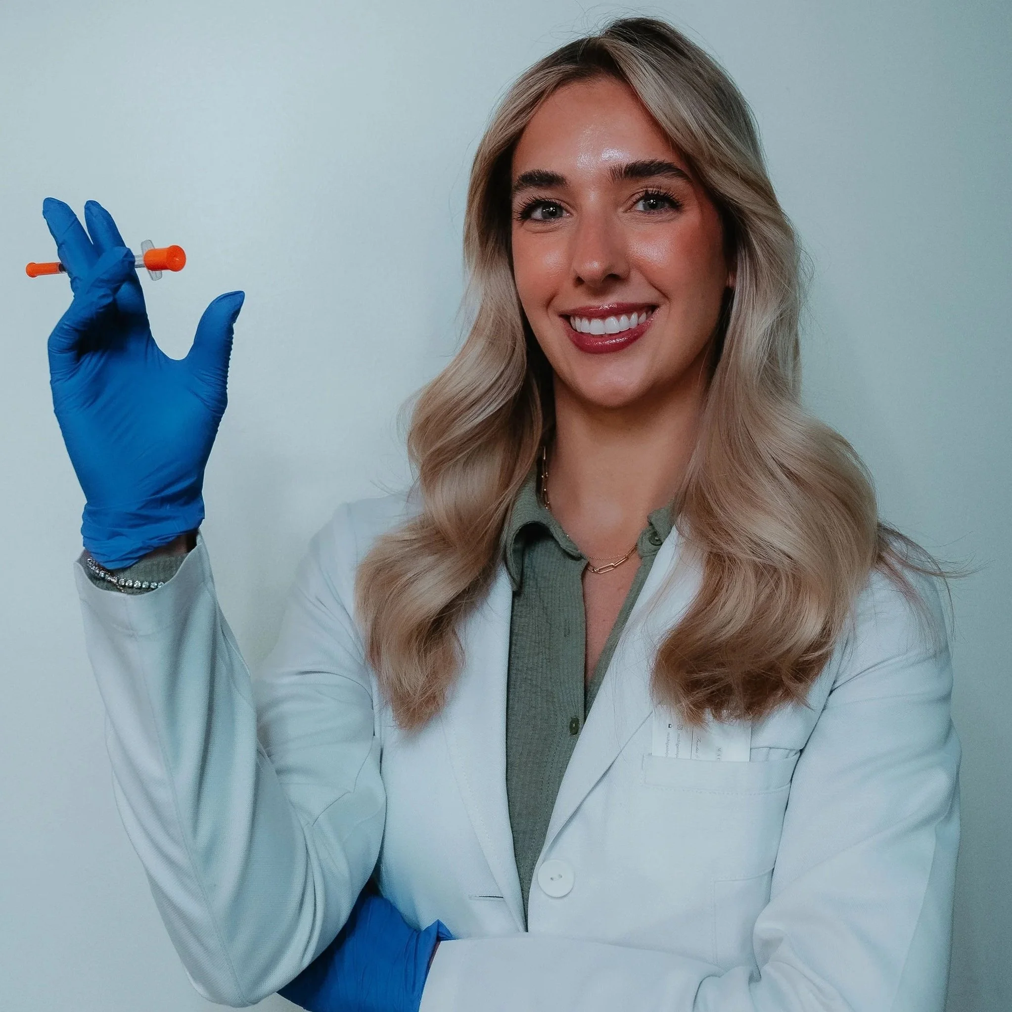 A smiling female healthcare professional wearing a white lab coat, green shirt, and blue gloves, holding a syringe.