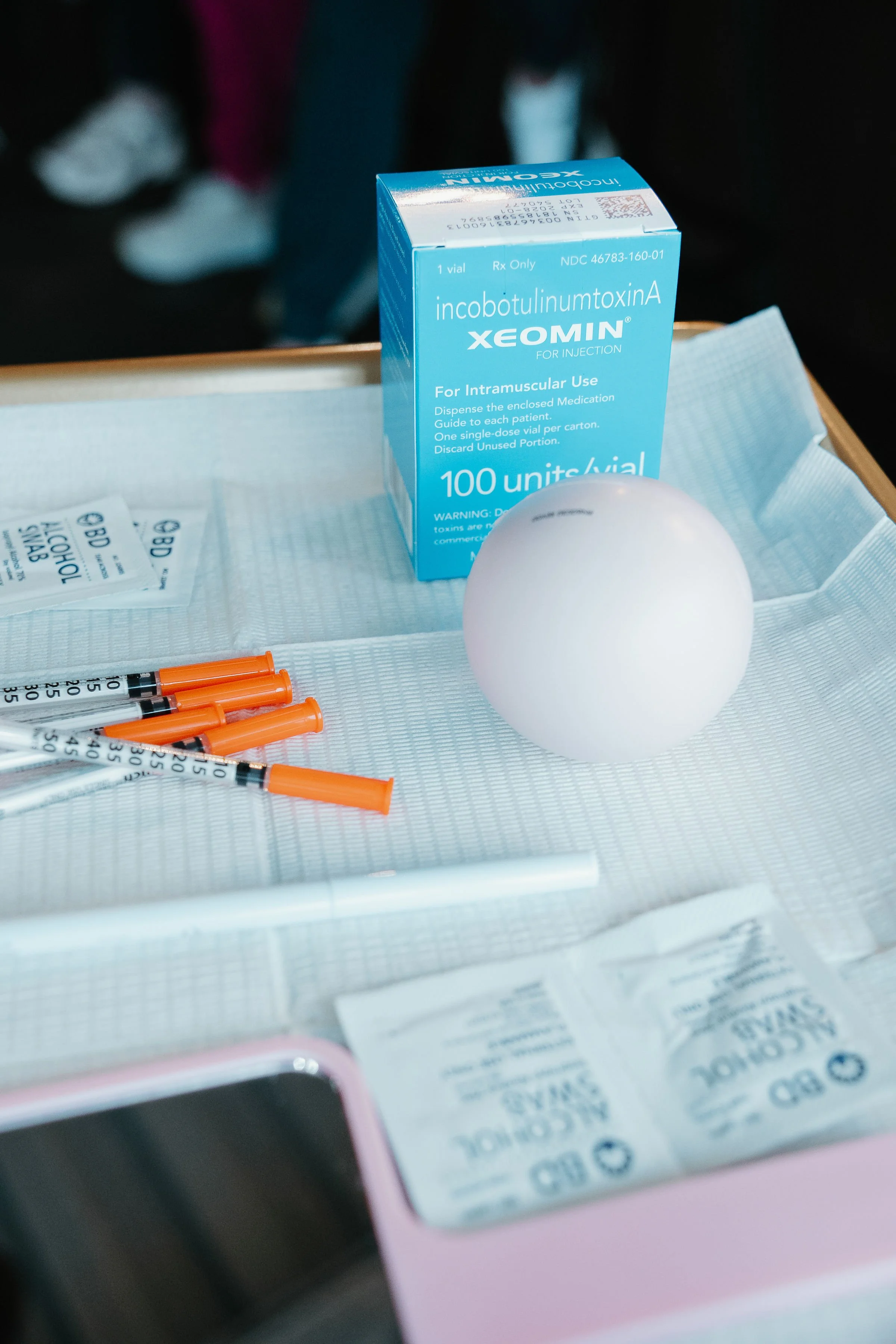 Medical supplies on a tray, including a box of Xeomin injectable medication, several syringes, a white foam ball, alcohol swabs, and a small pink container.