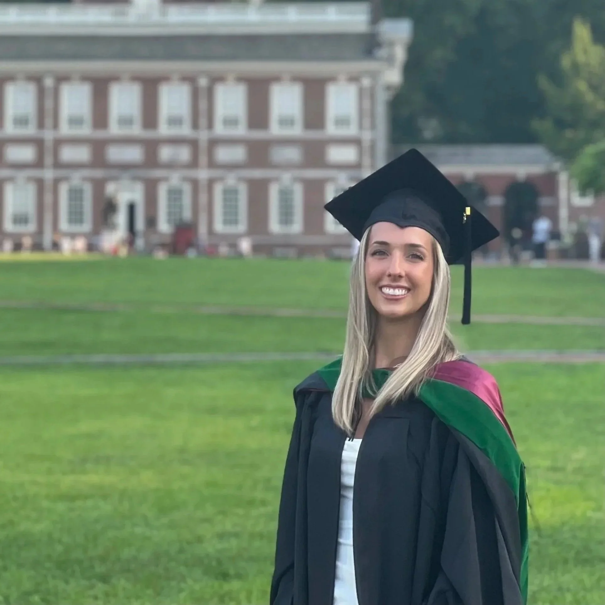 A young woman in a black graduation gown and cap, smiling, outdoors on a grassy area with a historic brick building in the background.