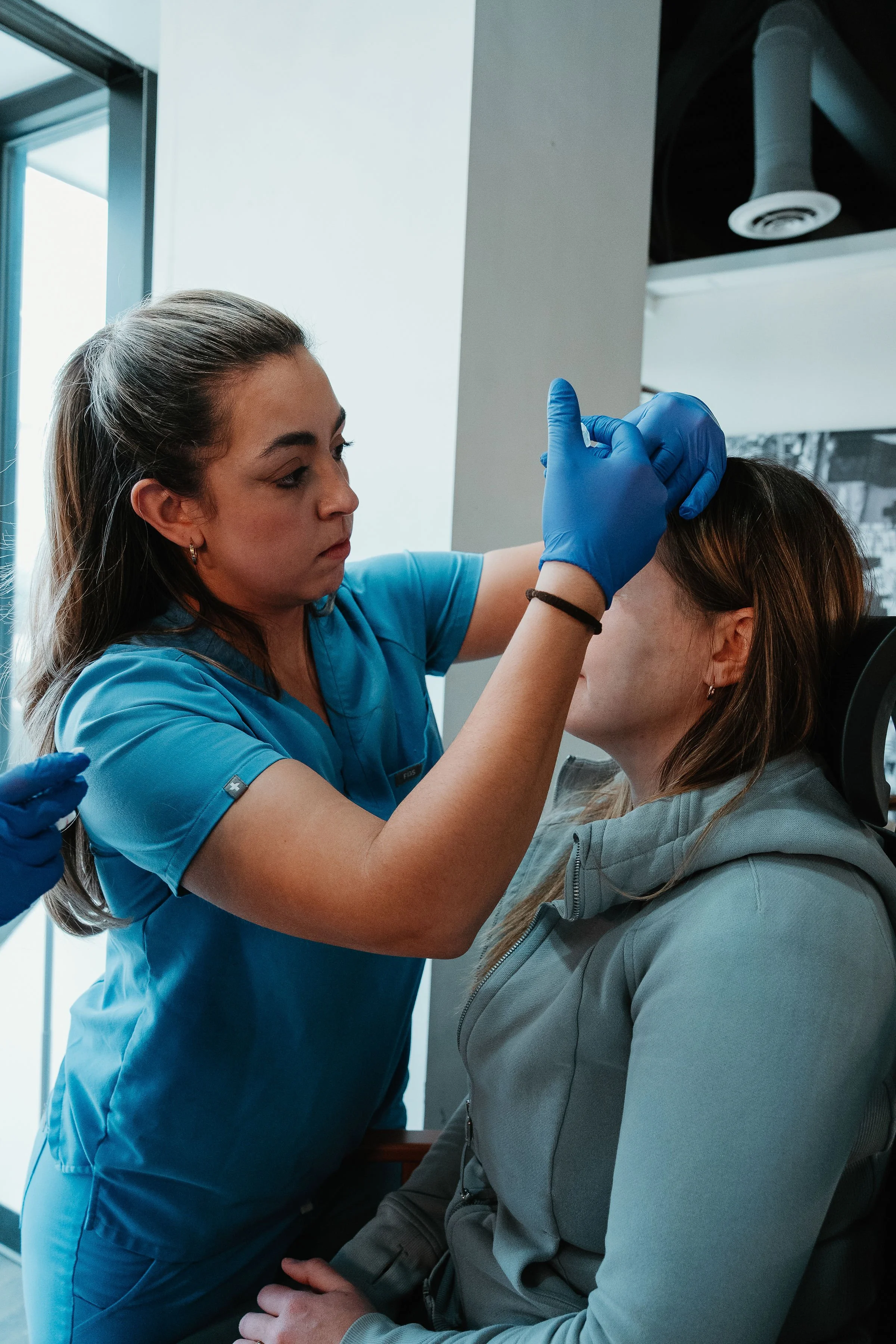A healthcare worker wearing blue scrubs and gloves administering a vaccine to a woman seated in a medical clinic.