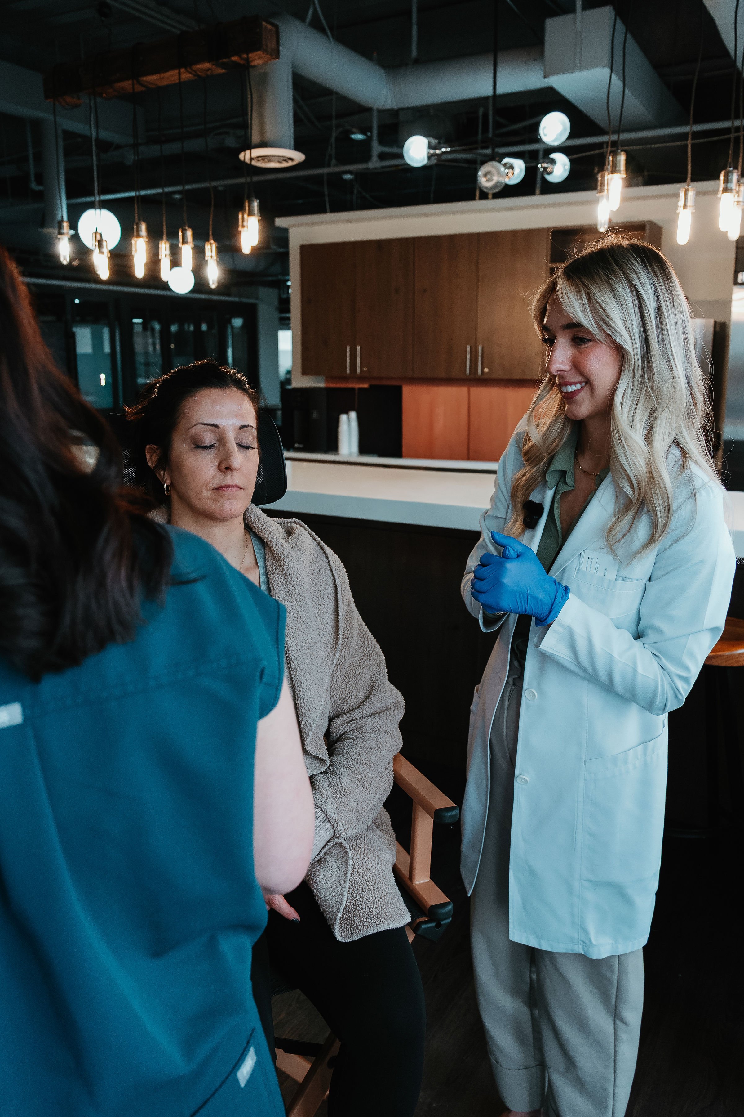 A woman in a gray fleece jacket sitting with her eyes closed having her arm prepared for a blood draw by a healthcare worker in dark scrubs, while another healthcare worker in a white coat and blue gloves looks on.