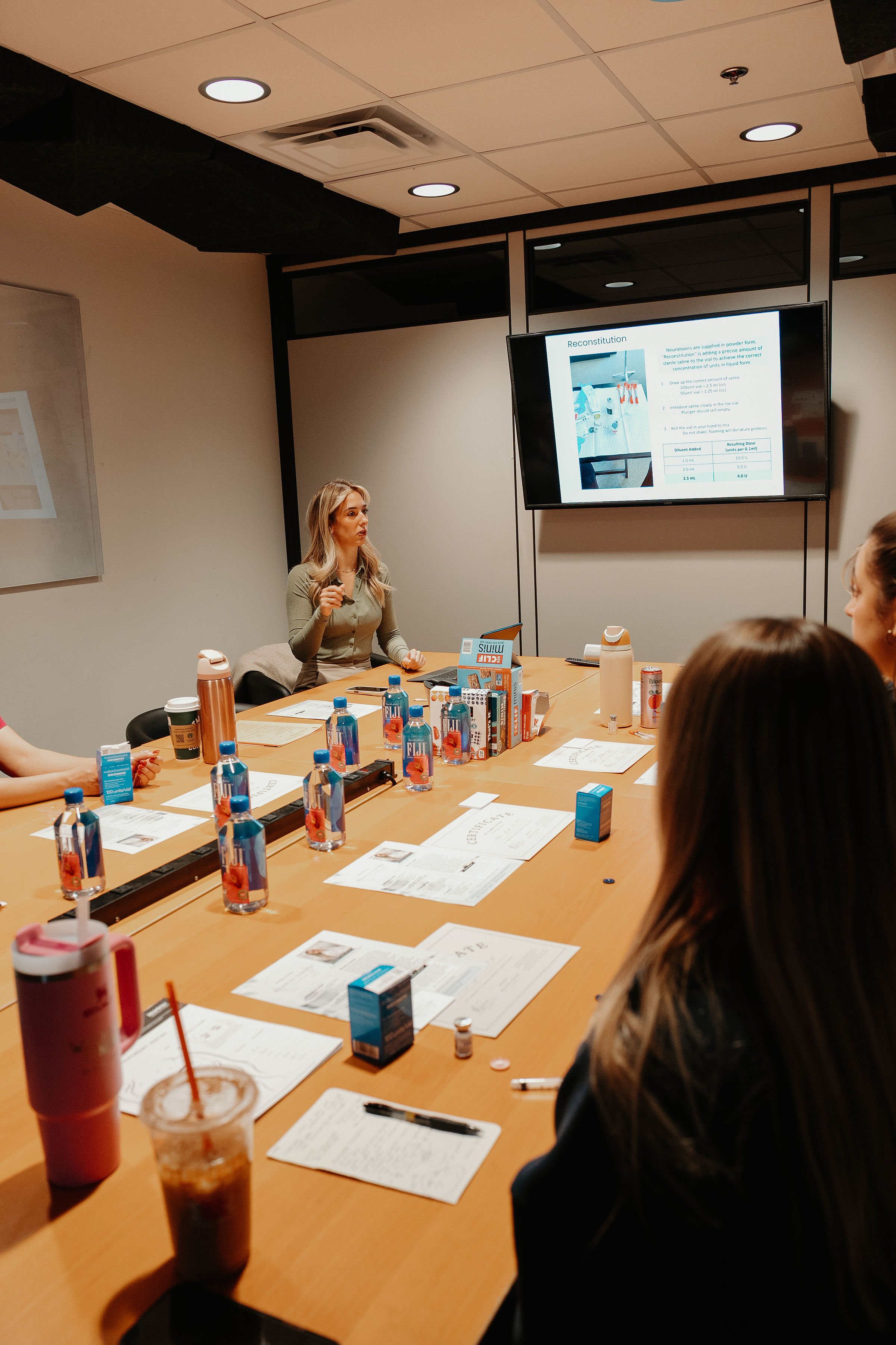 A woman giving a presentation in a conference room with a large screen displaying a slide. Several people are sitting around the table, with drinks, papers, and supplies on the table.