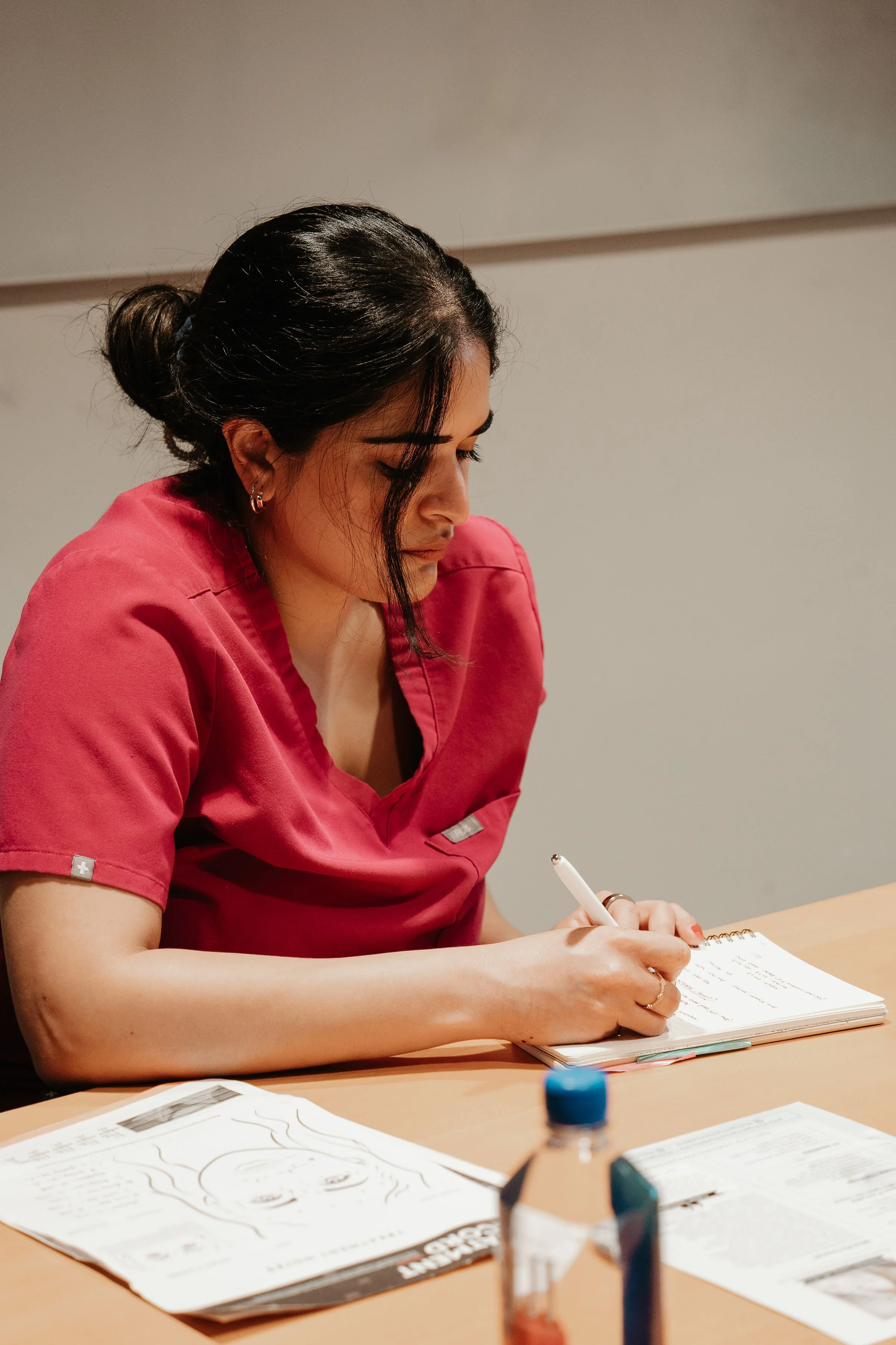 A woman in a pink medical uniform writing in a notebook at a desk, with a bottle and papers nearby.