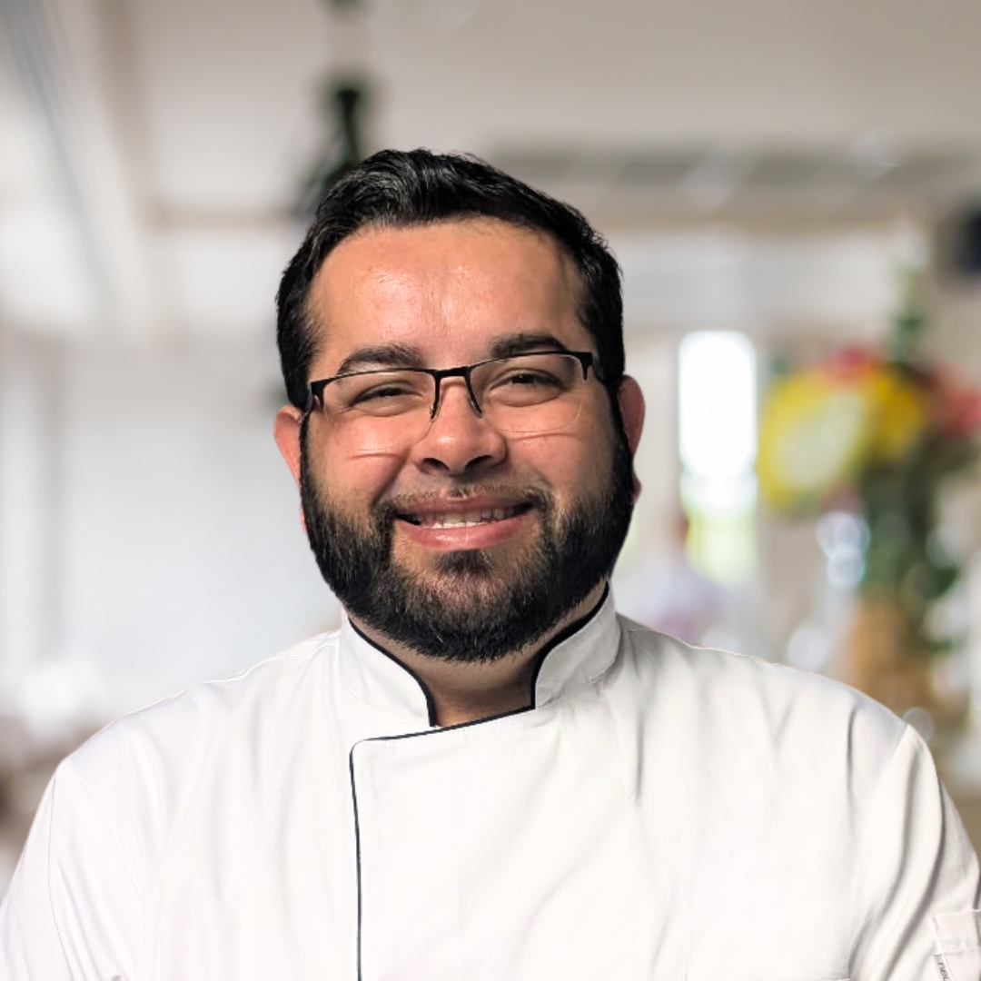 A smiling man with glasses and a beard in a chef's uniform in a bright kitchen.