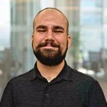 A smiling man with a beard and short hair, wearing a black collared shirt, standing in front of a blurred indoor background.