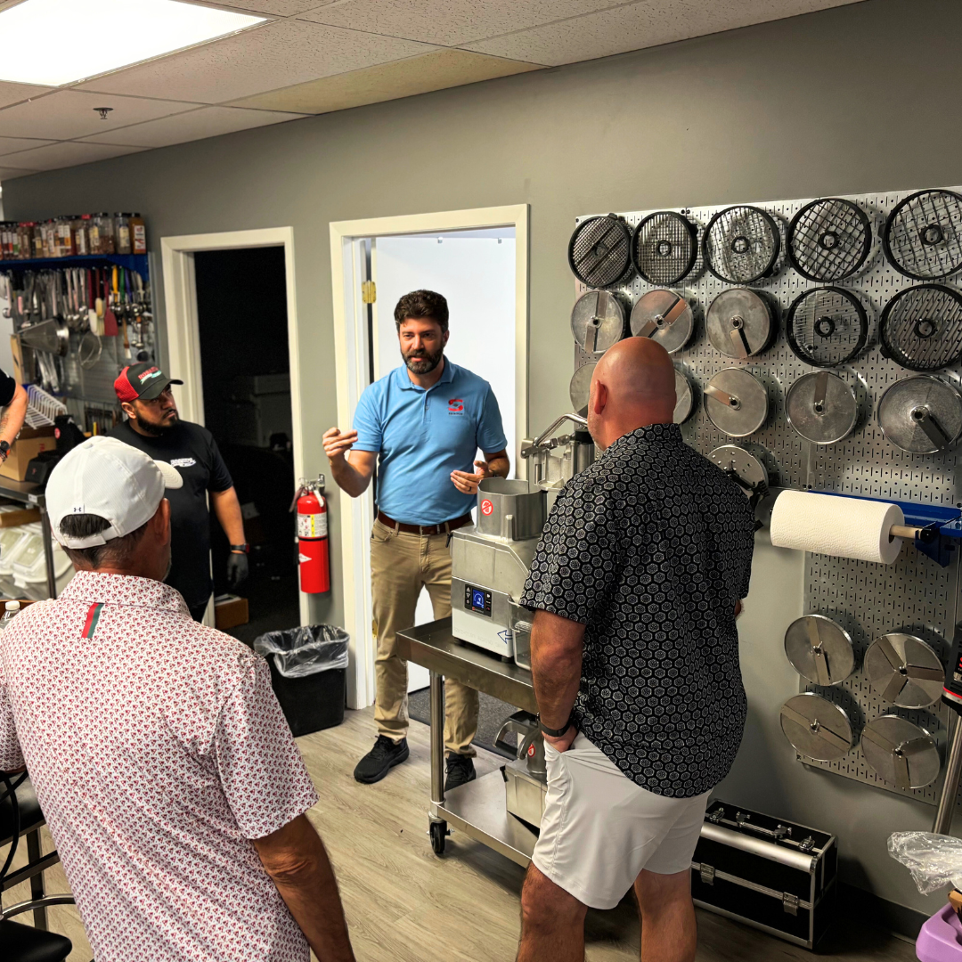 A group of five men having a discussion in a commercial kitchen or store. One man in a blue polo hosts, while others listen. Equipment and tools are visible on the walls, including a grinder and various grill plates.