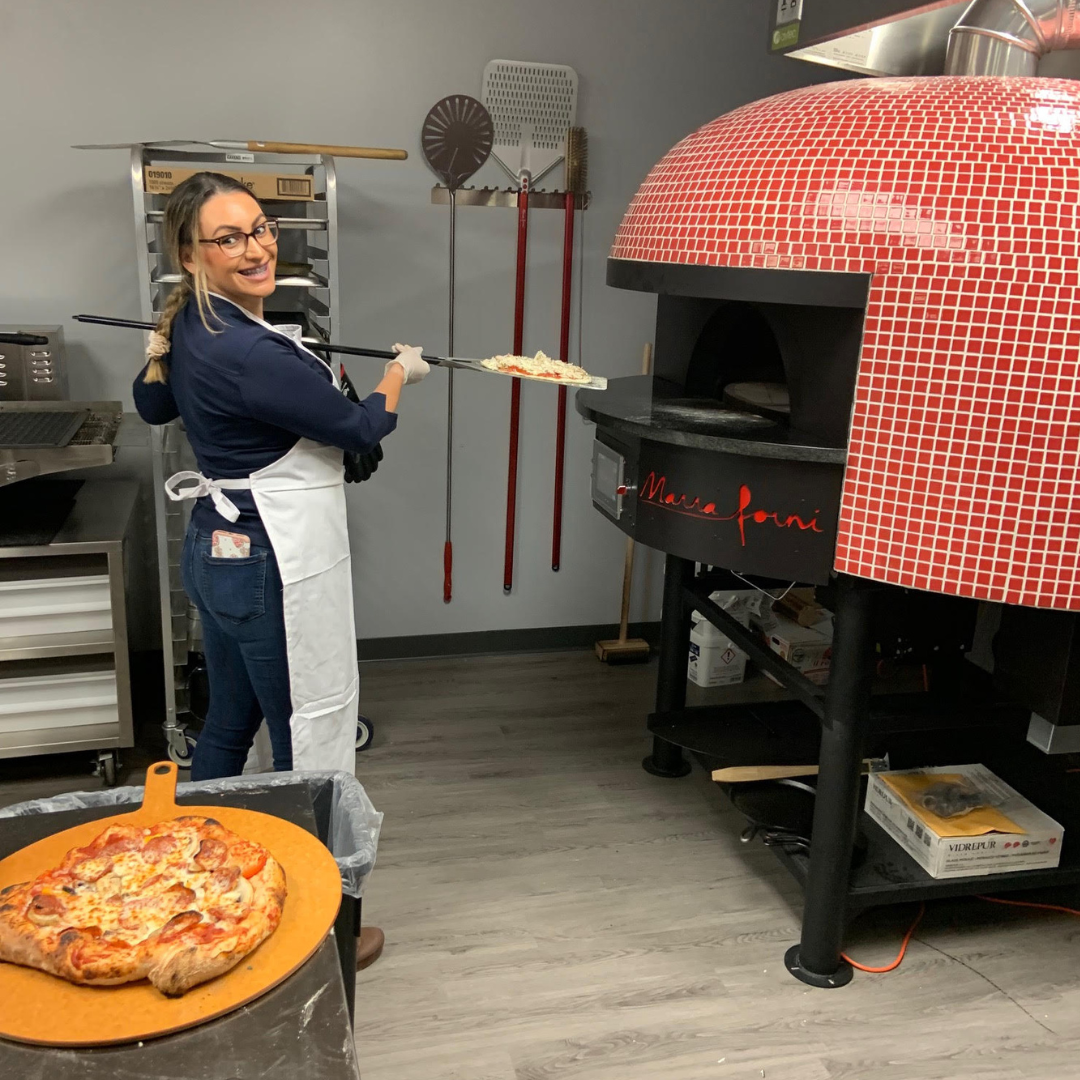 A woman in a white apron and glasses is smiling and holding a pizza peel with a pizza outside a wood-fired oven in a pizzeria kitchen.