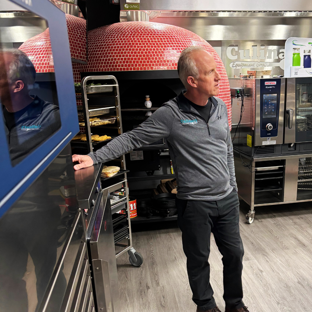 James Carr in the Culinary Equipment Group kitchen near a Marra Forni pizza oven and an Irinox MulitFresh with his right arm extended and hand resting on a stainless steel counter.