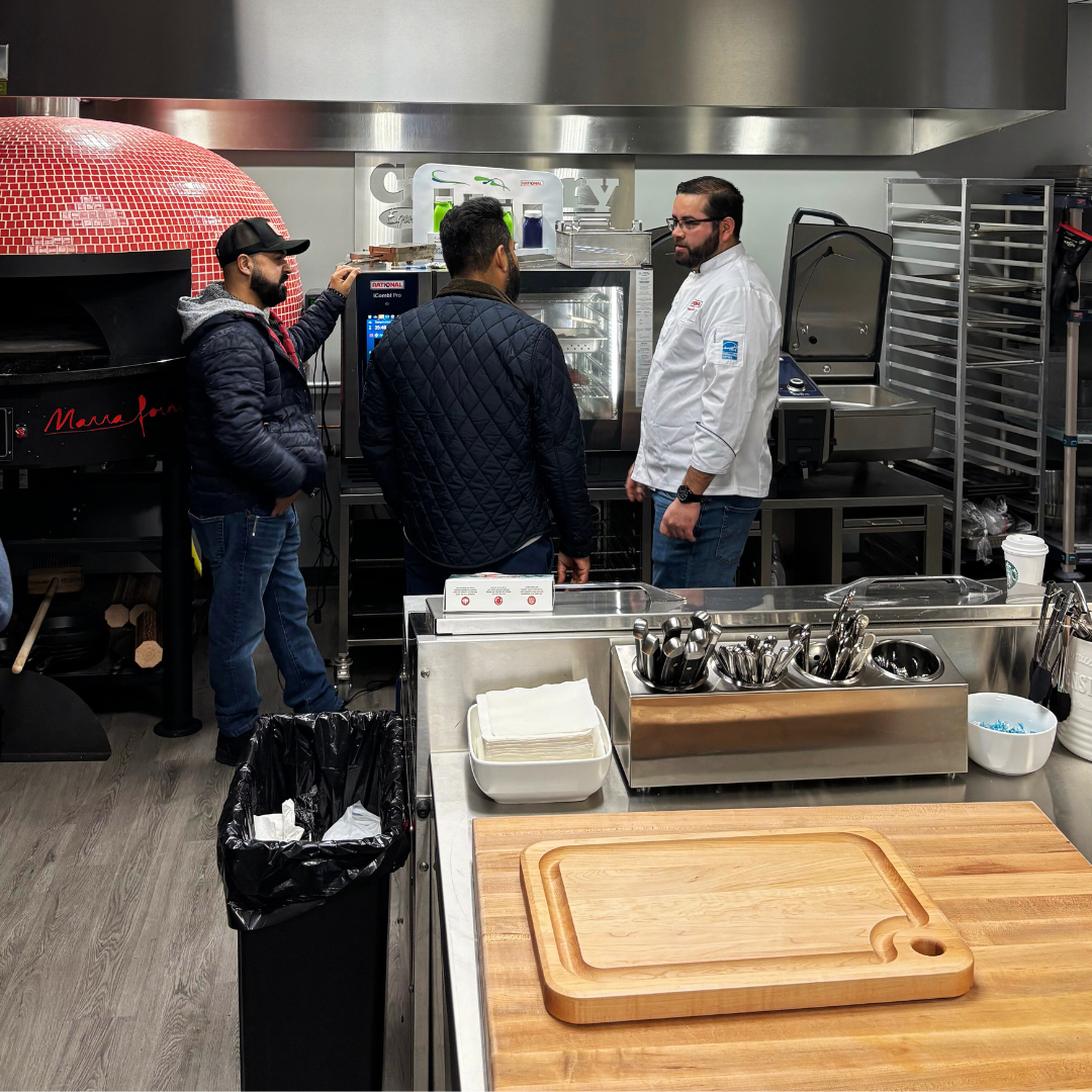 Three men having a conversation in a commercial kitchen, one is in a chef's coat, and the others are in casual clothing, near a microwave oven and kitchen equipment.