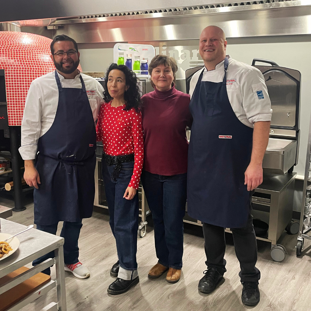 Four people, two men and two women, standing in a kitchen or restaurant setting, smiling at the camera. The men are wearing aprons, and the women are dressed casually. Behind them are kitchen equipment, including a pizza oven and a cart with bottles.