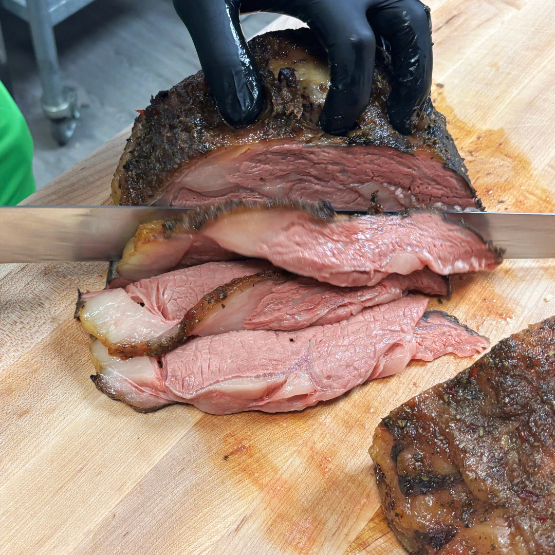  sliced medium-rare beef brisket on a wooden cutting board with a person wearing black gloves slicing it.