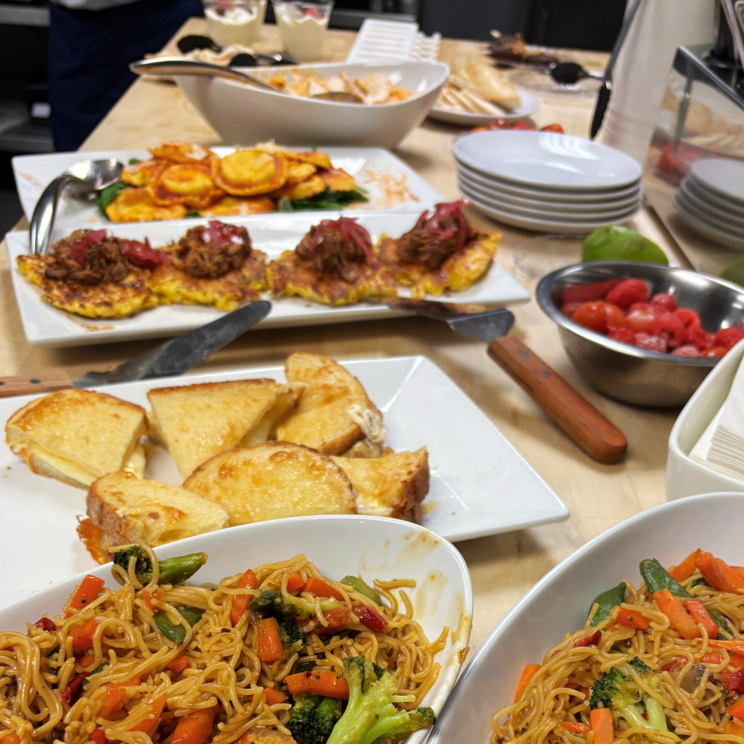 Variety of dishes including fried noodles with vegetables, garlic bread, pizza with toppings, and a bowl of berries on a wooden table