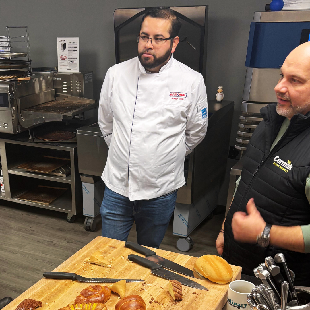Two men in a kitchen or bakery setting, one in a white chef's coat and the other in a black vest, standing near a wooden table with baked goods and knives.