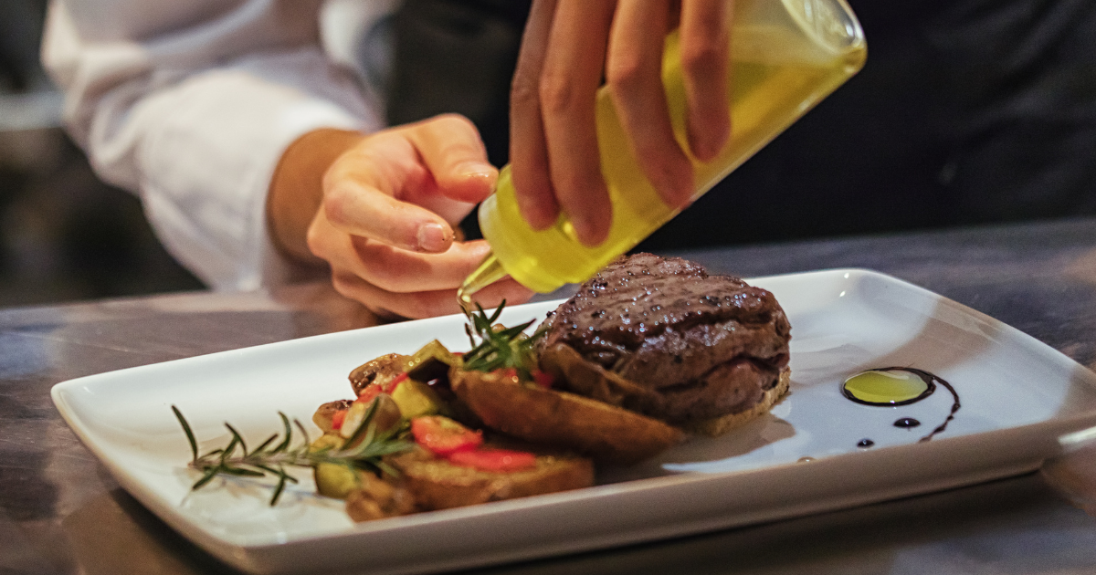 Person pouring olive oil over a steak with roasted vegetables on a white plate.