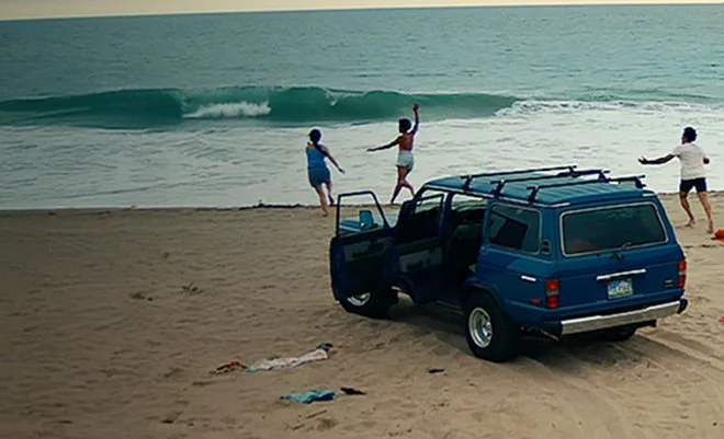 People playing on the beach near a blue SUV with an open door.