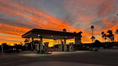 A gas station at sunset with orange and pink clouds, palm trees in the background, and multiple fueling pumps in the foreground.