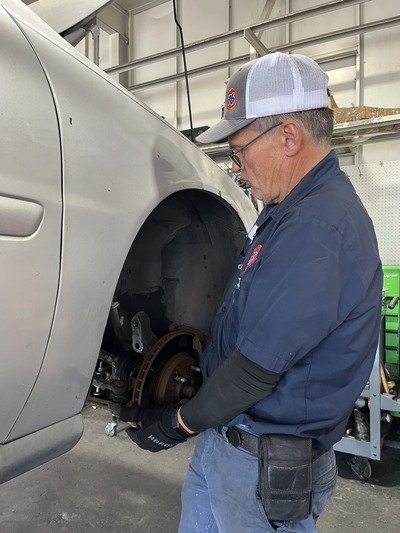 A man wearing a baseball cap and glasses working on a car in a garage or workshop, with the car's front wheel removed.