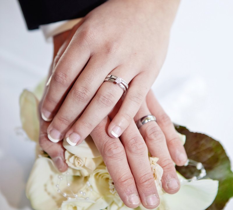 A woman's and a man's hand wearing wedding rings rest on top of a bouquet of white flowers.