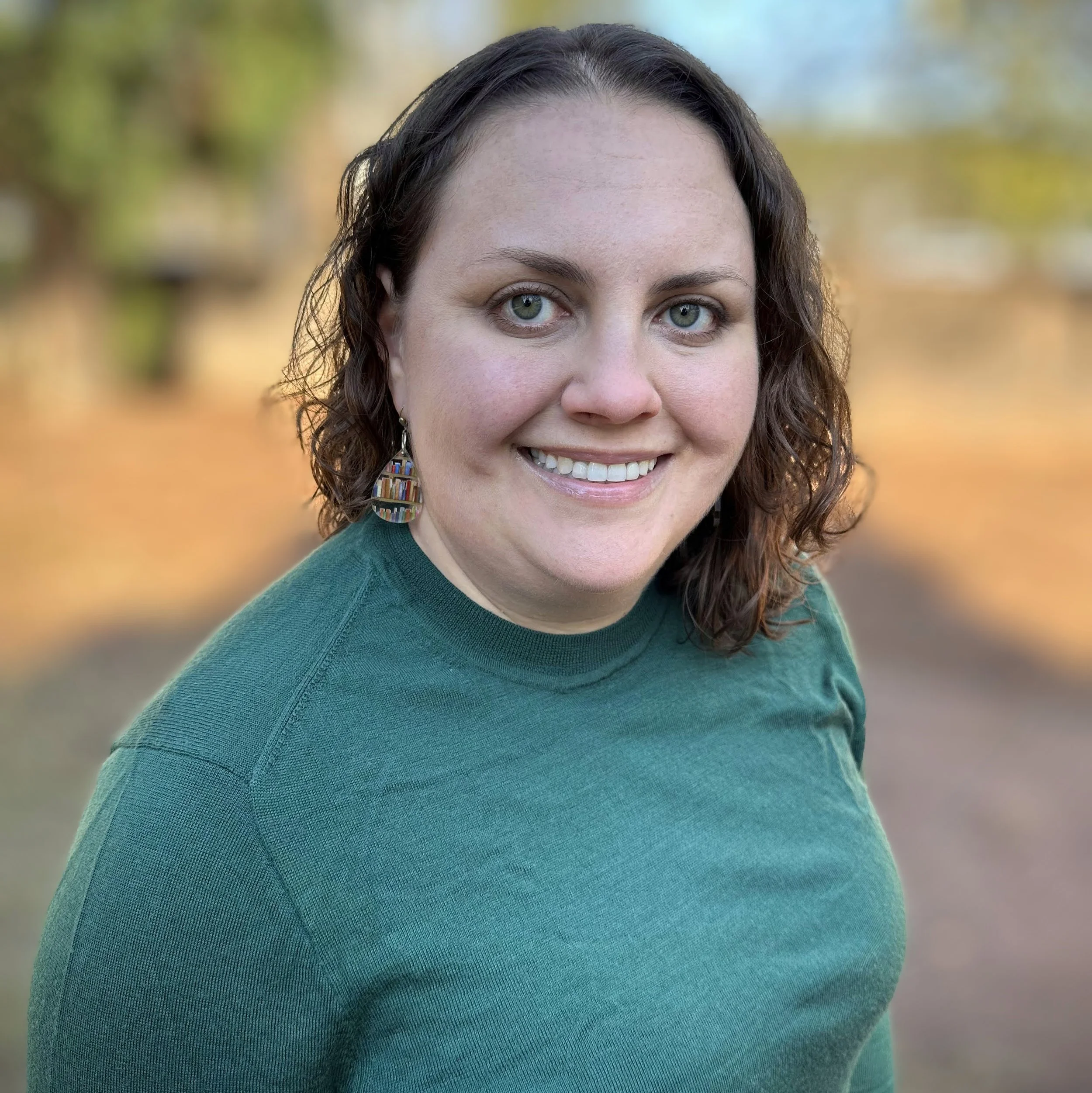 A woman with curly brown hair and green eyes, wearing a green sweater and drop earrings with books on them, smiles at the camera.