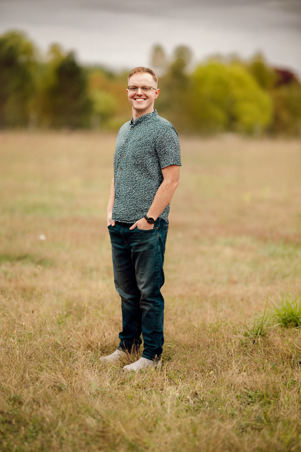 A young man stands in a field with trees in the background, smiling at the camera. He is wearing glasses, a patterned short-sleeve shirt, dark jeans, and white socks.