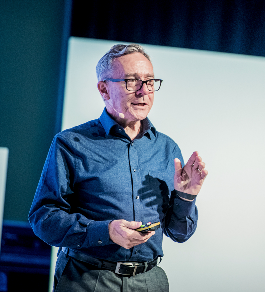A man wearing glasses and a blue button-up shirt giving a presentation with a wireless microphone headset, holding a remote control, in front of a white screen.
