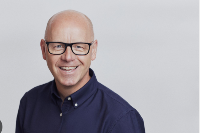 Smiling bald man with glasses wearing a navy blue button-up shirt against a light gray background.