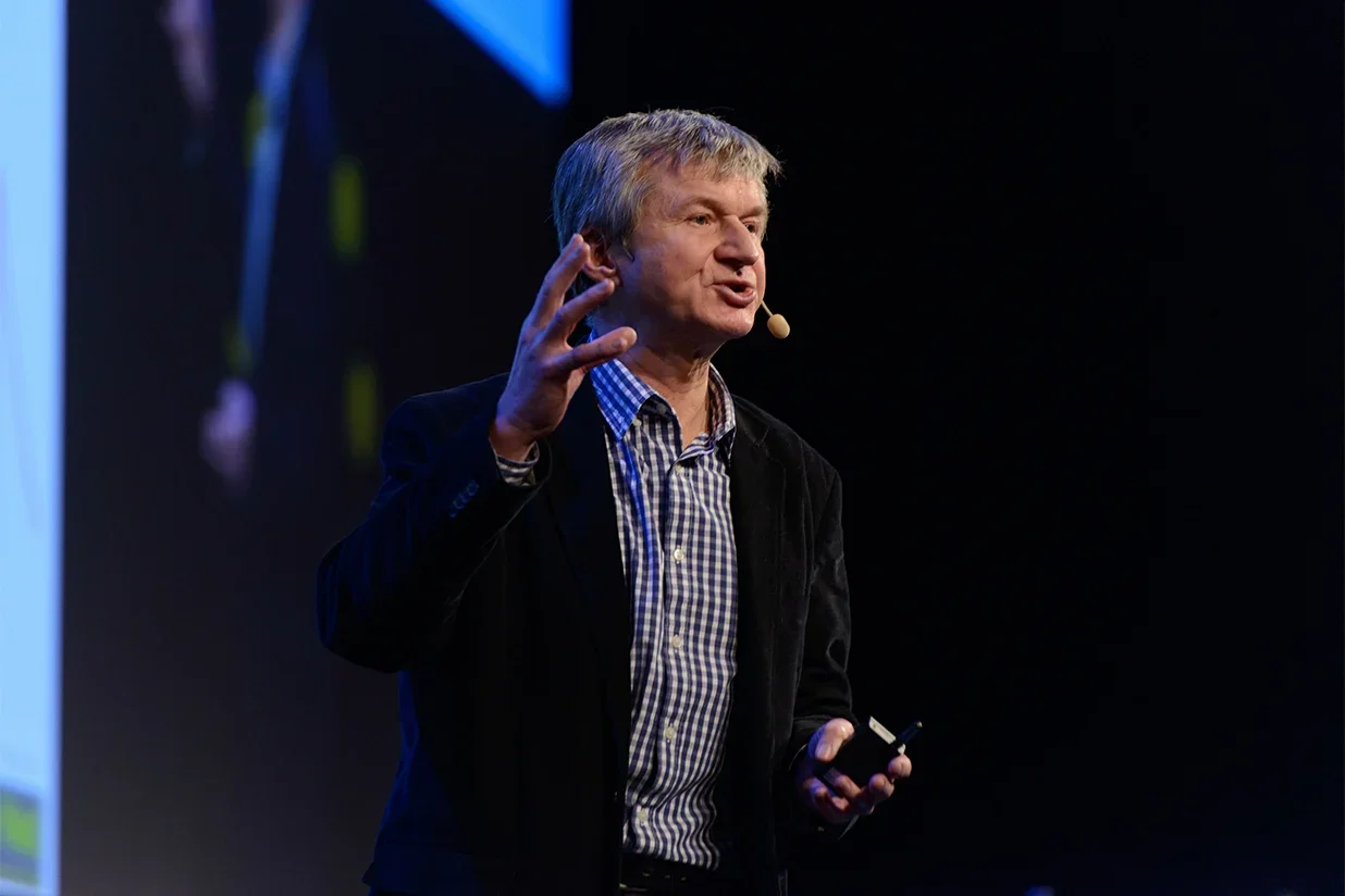 A man with gray hair speaking on a stage, wearing a checkered shirt and a dark blazer, holding a microphone near his face and gesturing with his left hand.