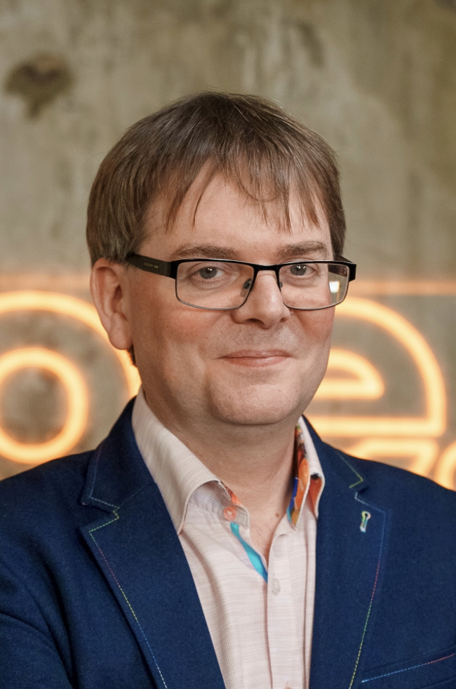 A man with glasses wearing a dark blue blazer and a light shirt, standing in front of a blurred sign with orange neon lights.